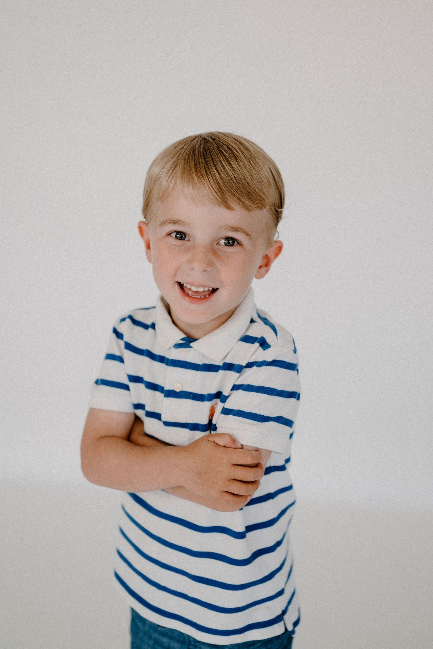 Happy blonde boy in blue striped polo shirt smiling with arms crossed in white studio.