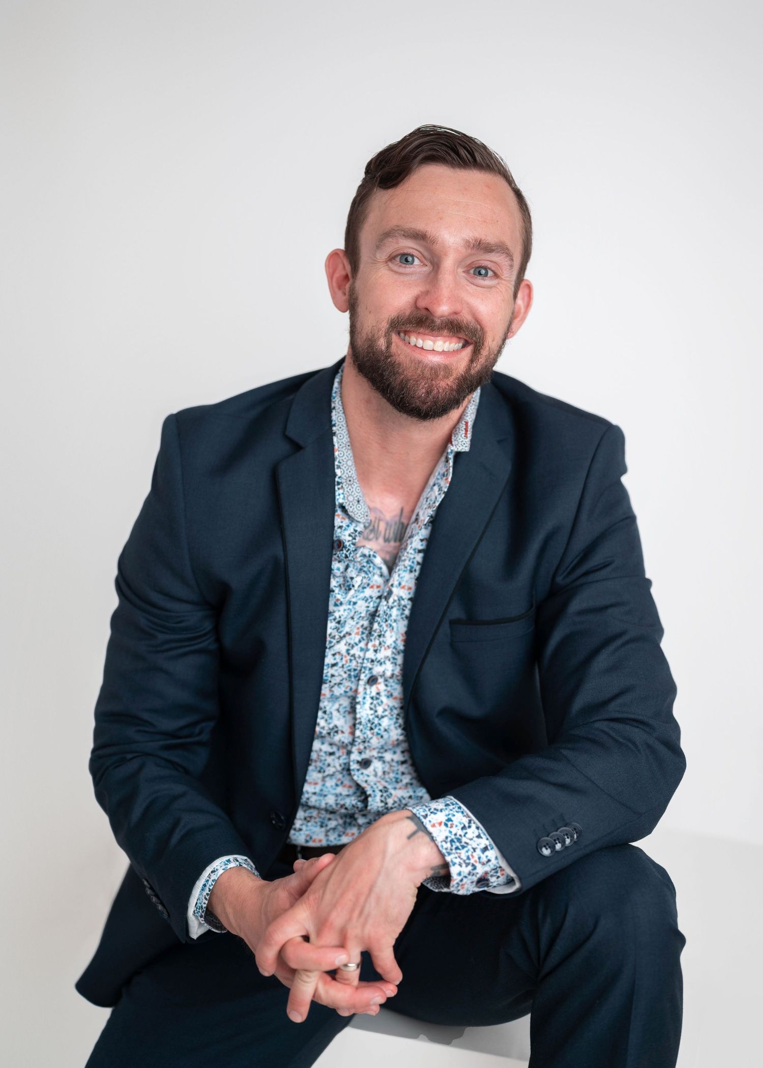 Smiling bearded man in navy suit and floral shirt sitting confidently against white background.