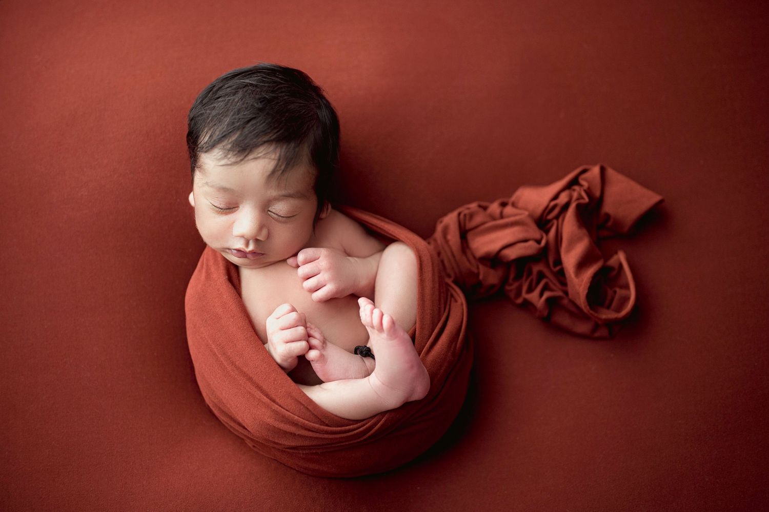 Newborn baby boy wrapped in rust-red swaddle sleeping peacefully on matching warm-toned studio backdrop.