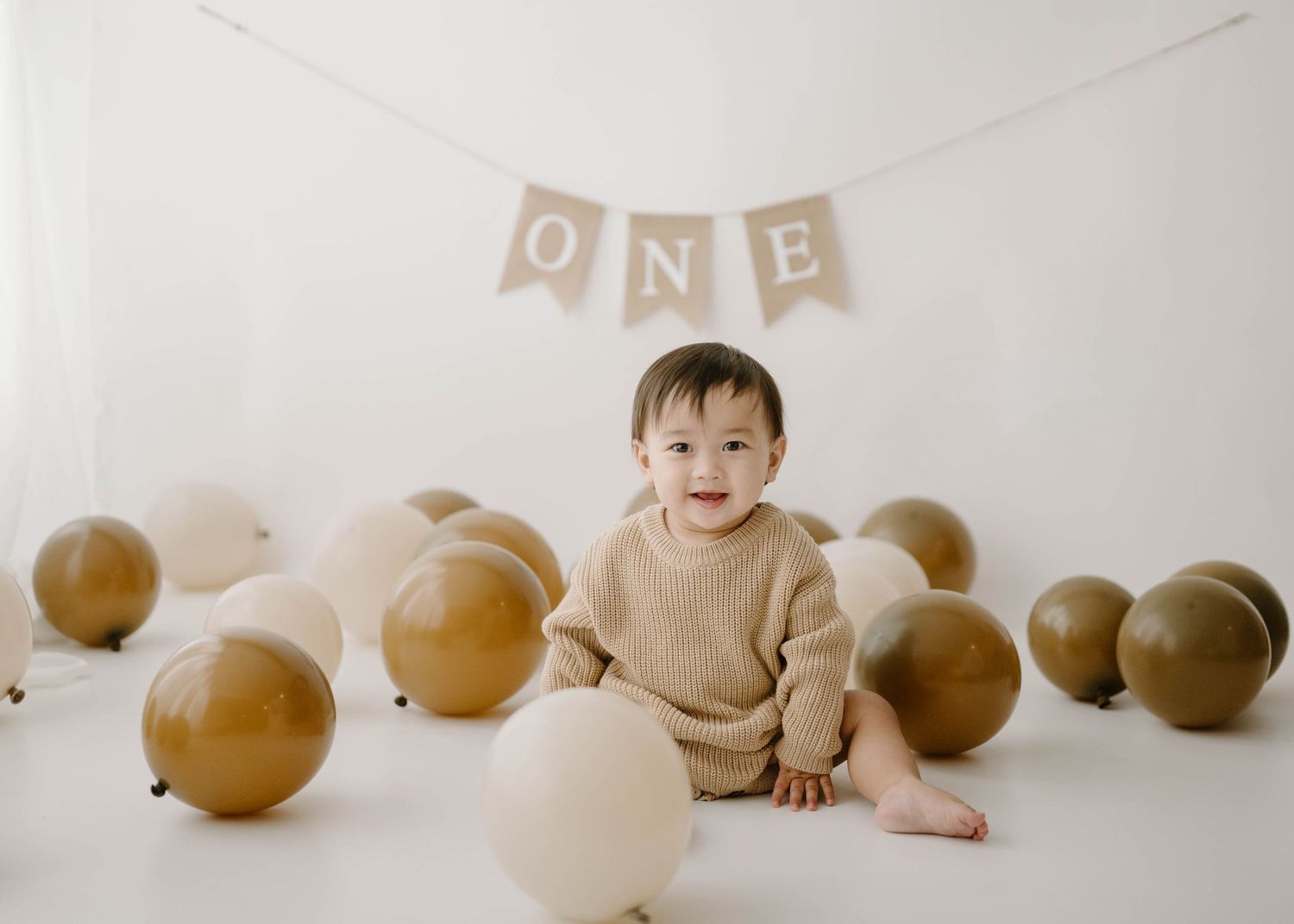A smiling baby in a beige knit outfit sits among brown and cream balloons under a burlap ONE banner for a first birthday.