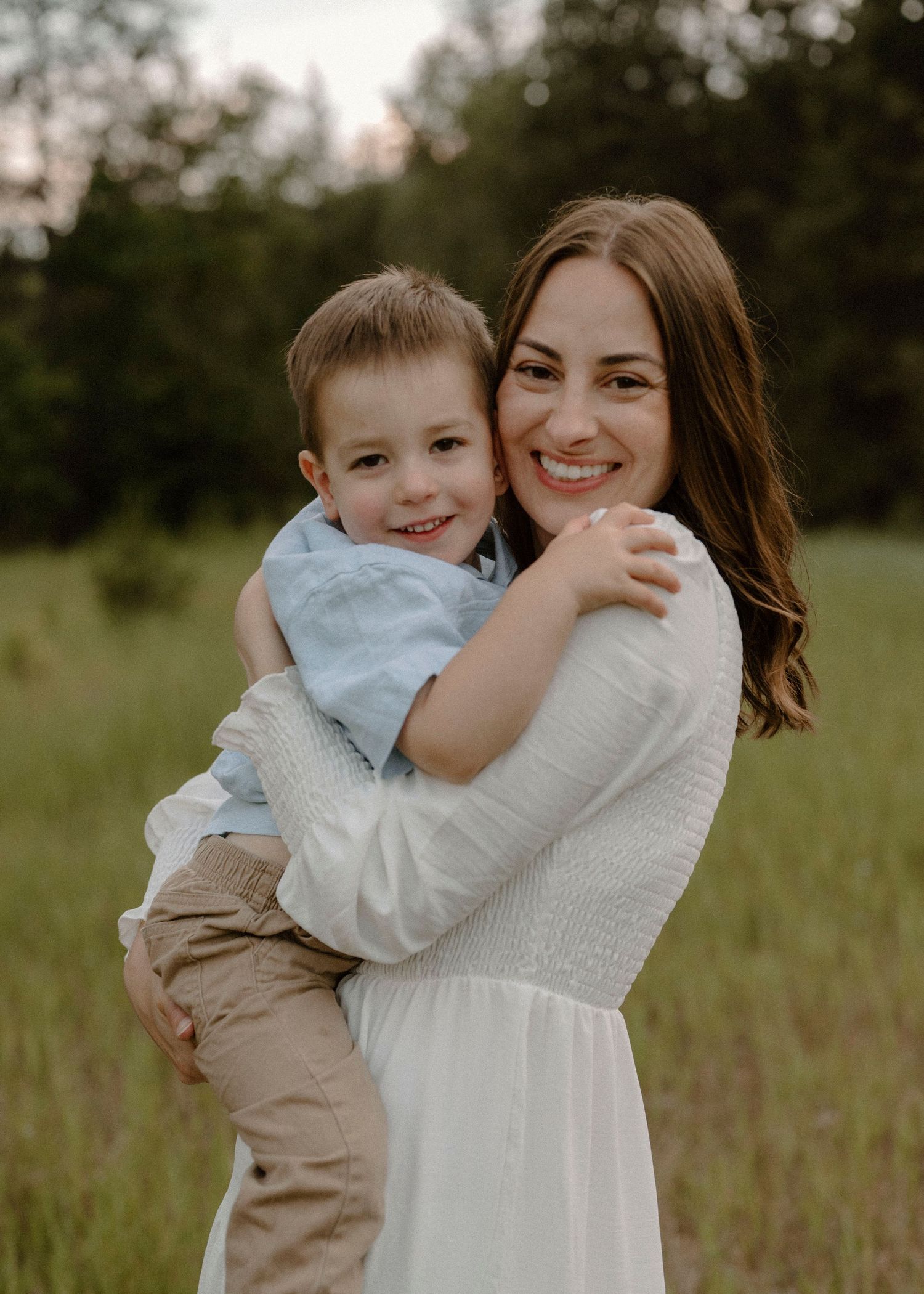 A smiling mother holds her young son in a sunlit green meadow.