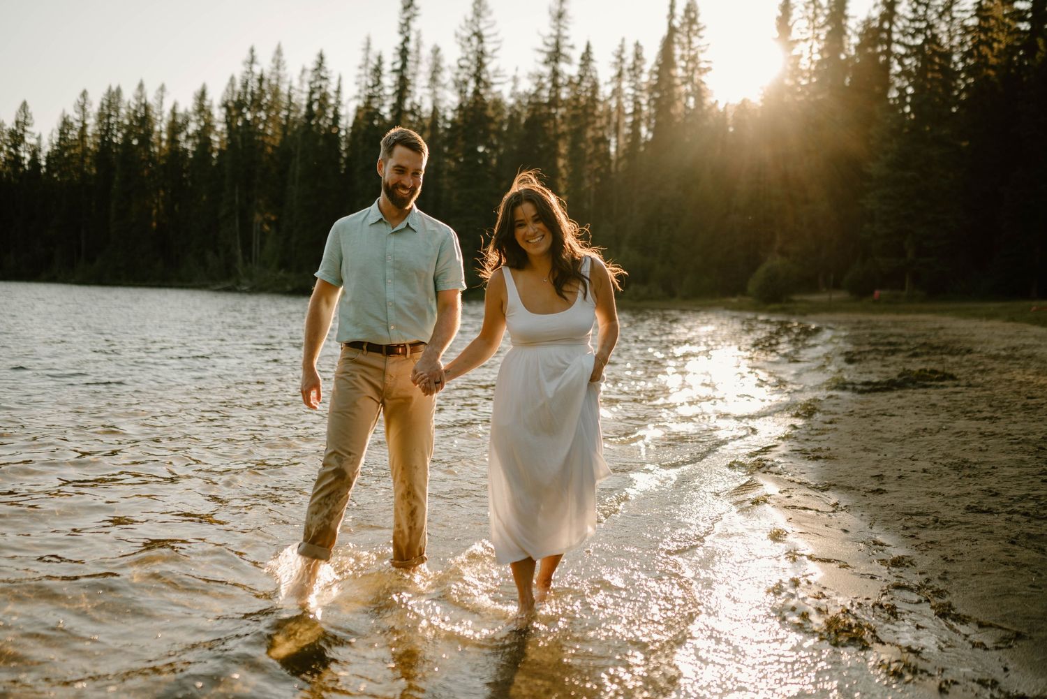 Couple holding hands and wading through a lake at golden hour with a pine forest backdrop.