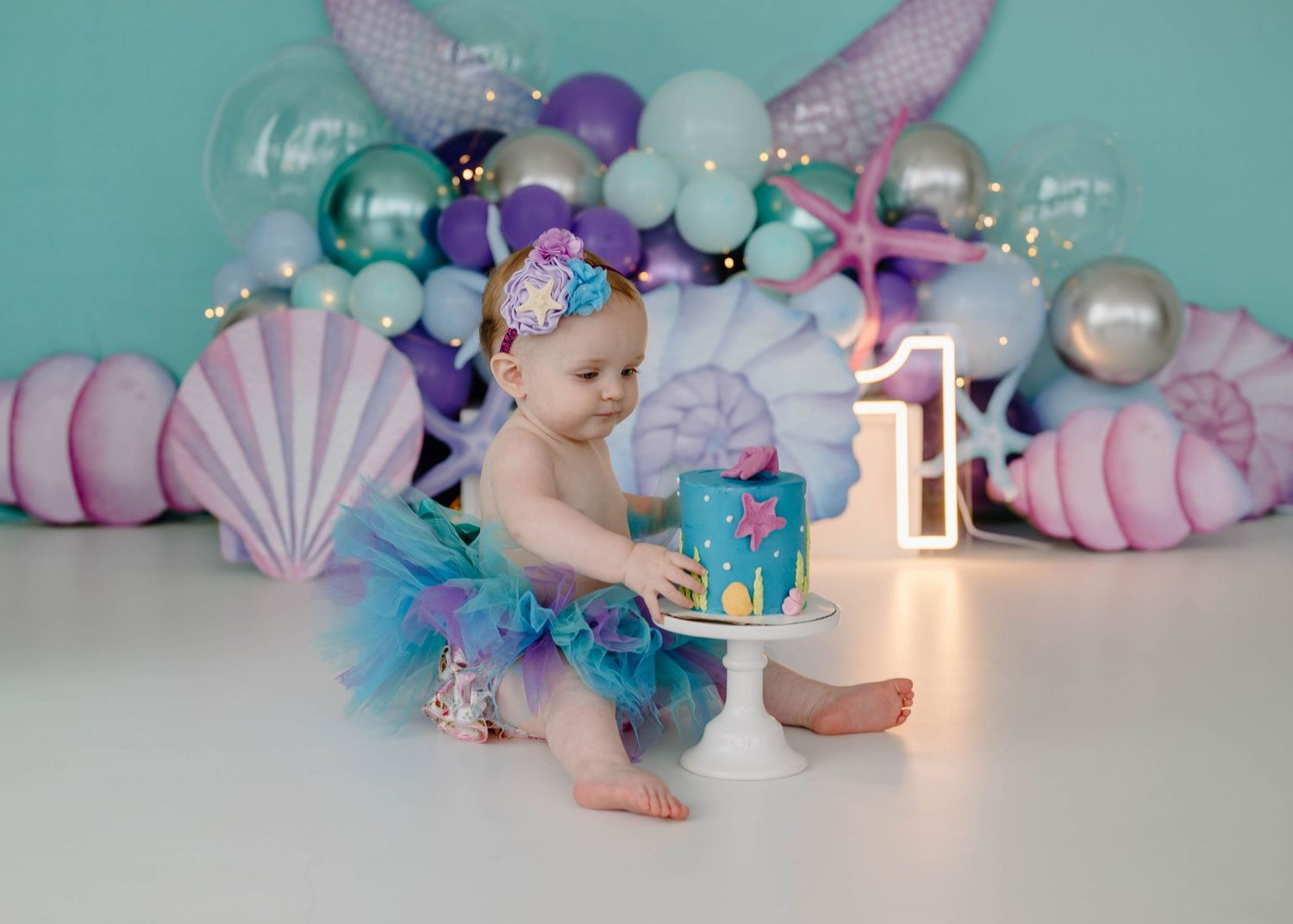 Baby girl in mermaid tutu smashes first birthday cake surrounded by ocean-themed balloon backdrop and seashell decorations.