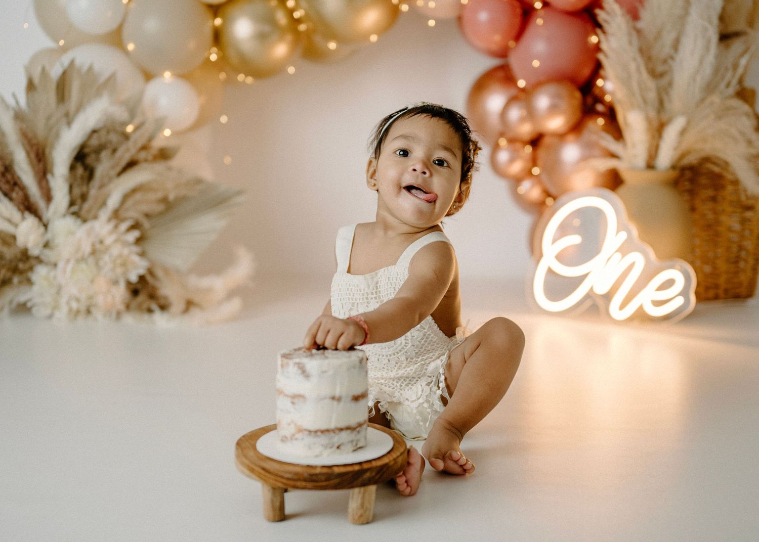 Baby girl smashes white cake at boho first birthday session with pink and gold balloon garland backdrop.