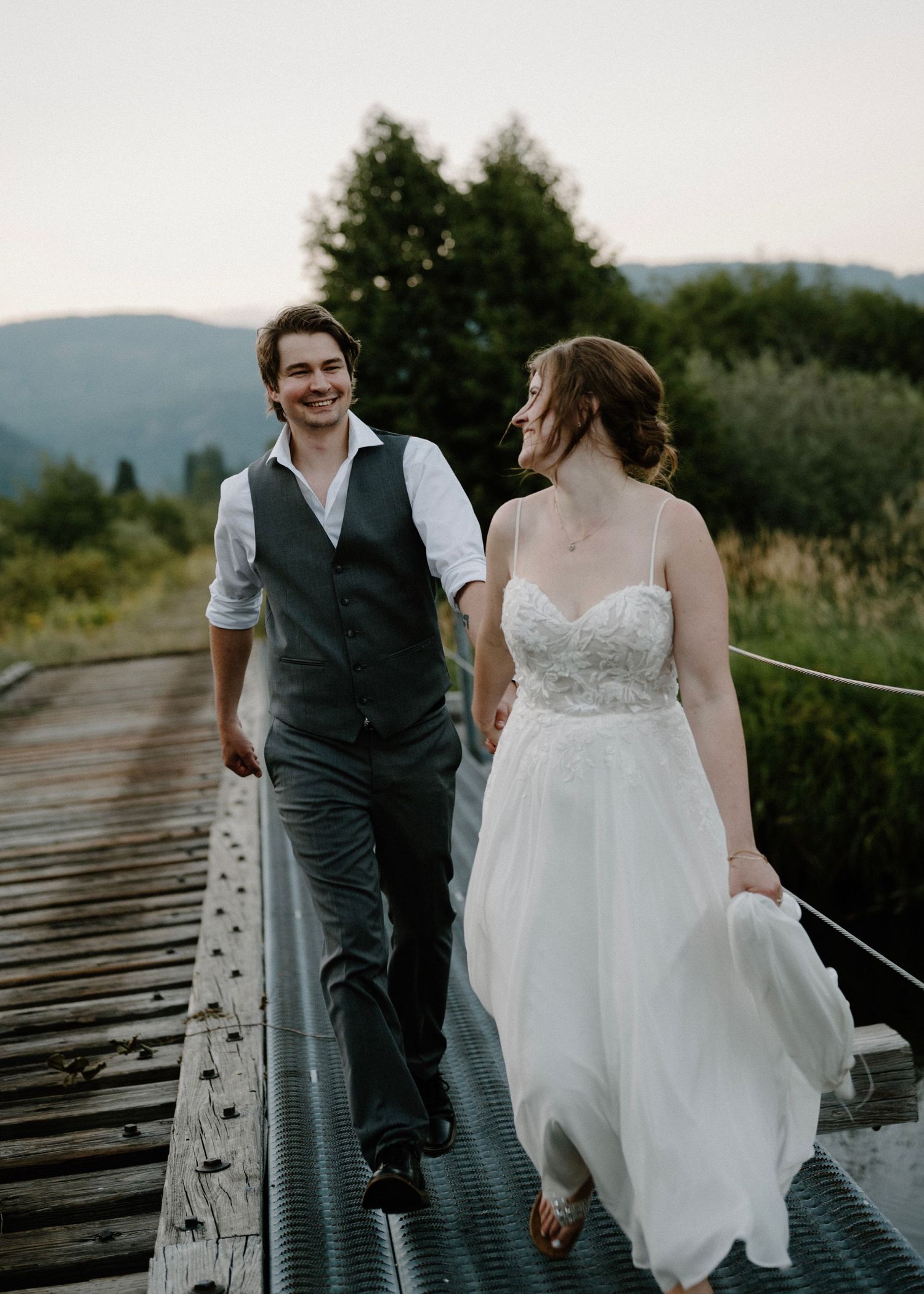 Bride and groom walking hand in hand on a bridge with scenic mountain views in the background.