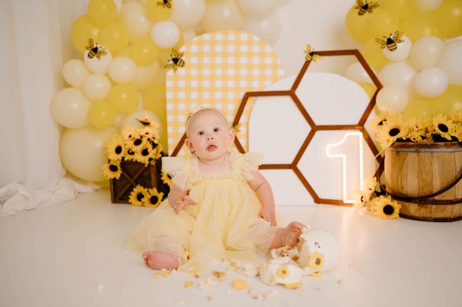 Baby girl in yellow dress sits by honeycomb decor, sunflowers, and balloons for a bee-themed first birthday cake smash.