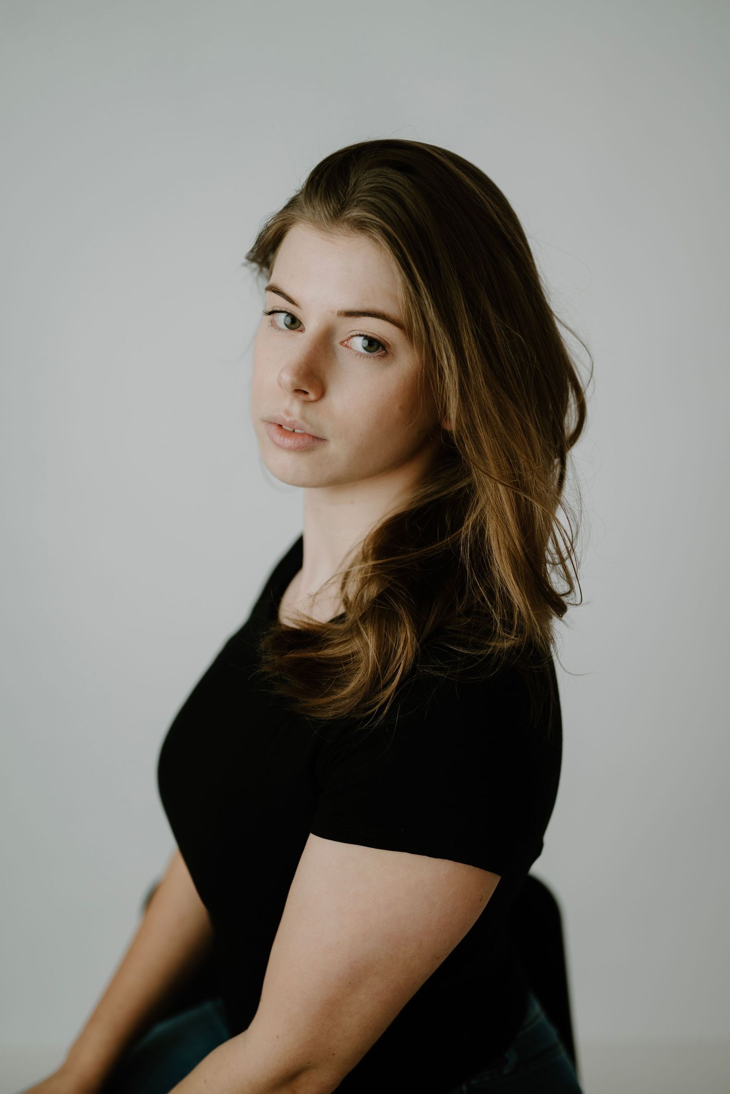Young woman with brown hair in a black top poses in a three-quarter turn for a professional portrait photo.