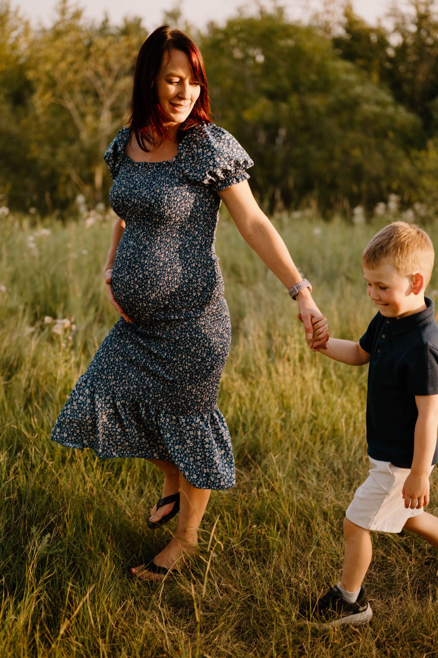 Pregnant redhead mother in floral dress holding hands with young blond son in field.