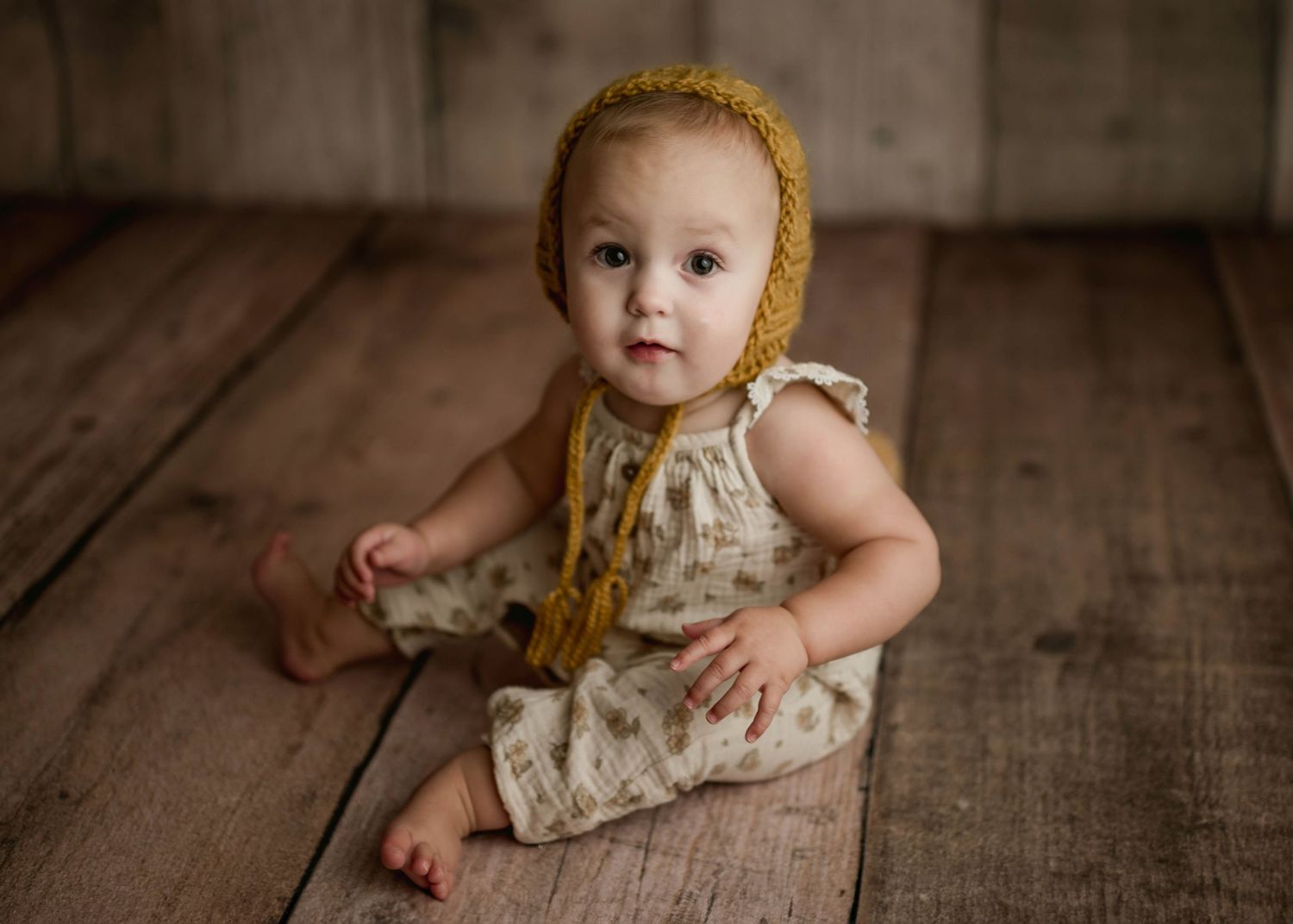 Baby girl in floral romper and golden yellow knit bonnet sits on rustic wood floor in studio.