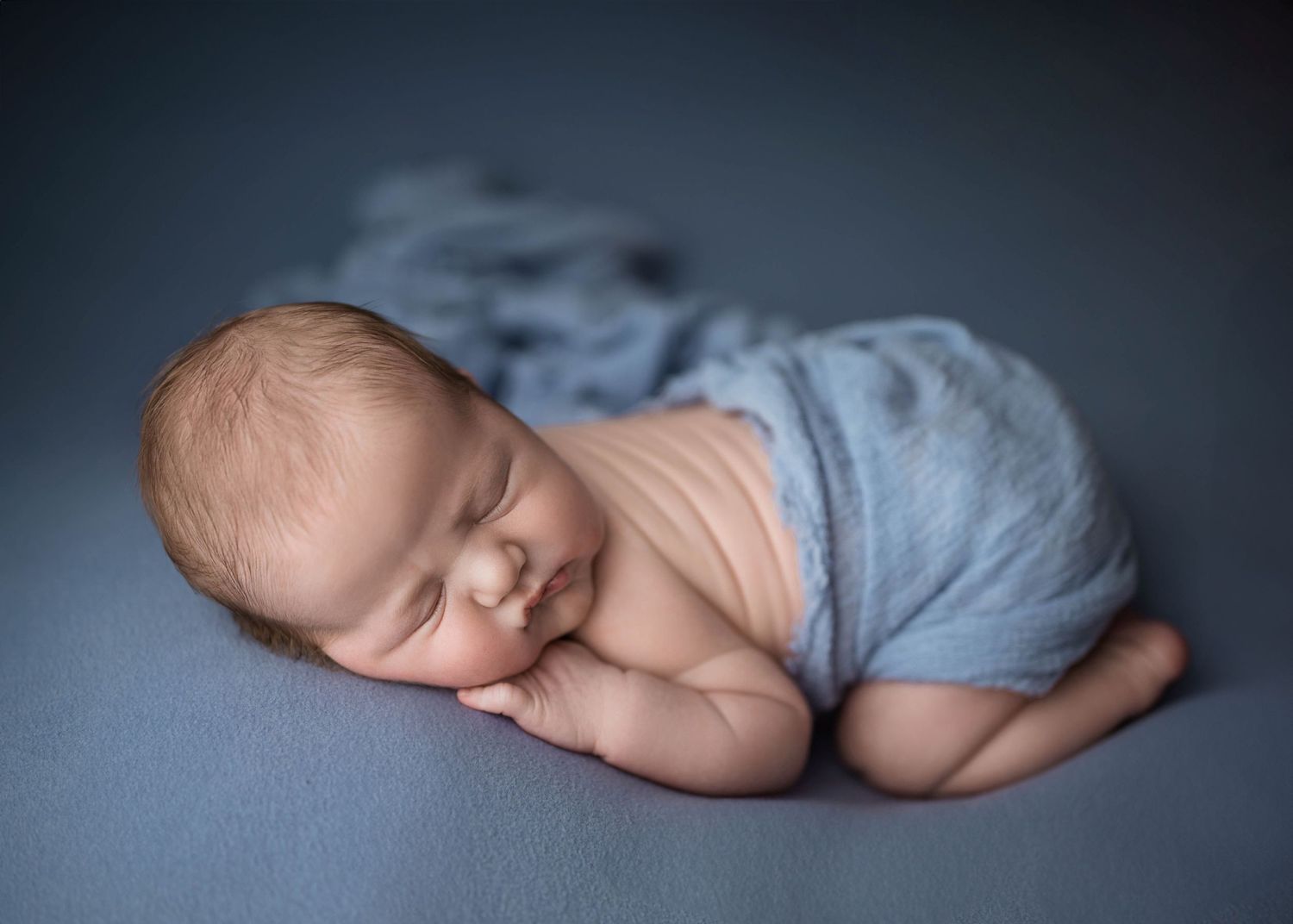 A peaceful newborn baby sleeping wrapped in a soft blue knit blanket on a gray backdrop.