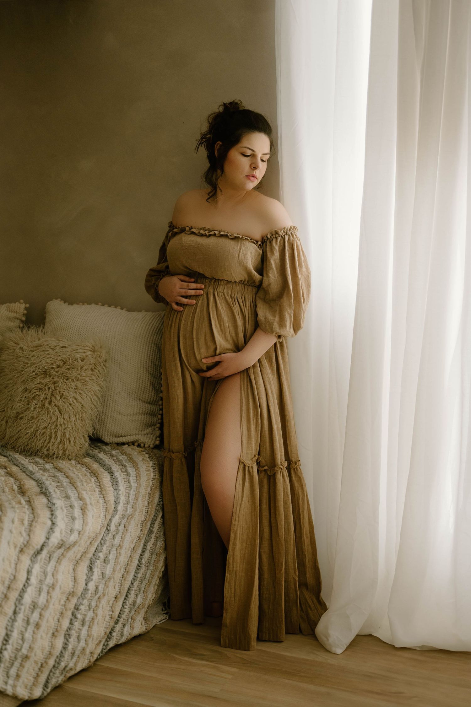 Pregnant woman in golden off-shoulder gown standing by window holding belly.