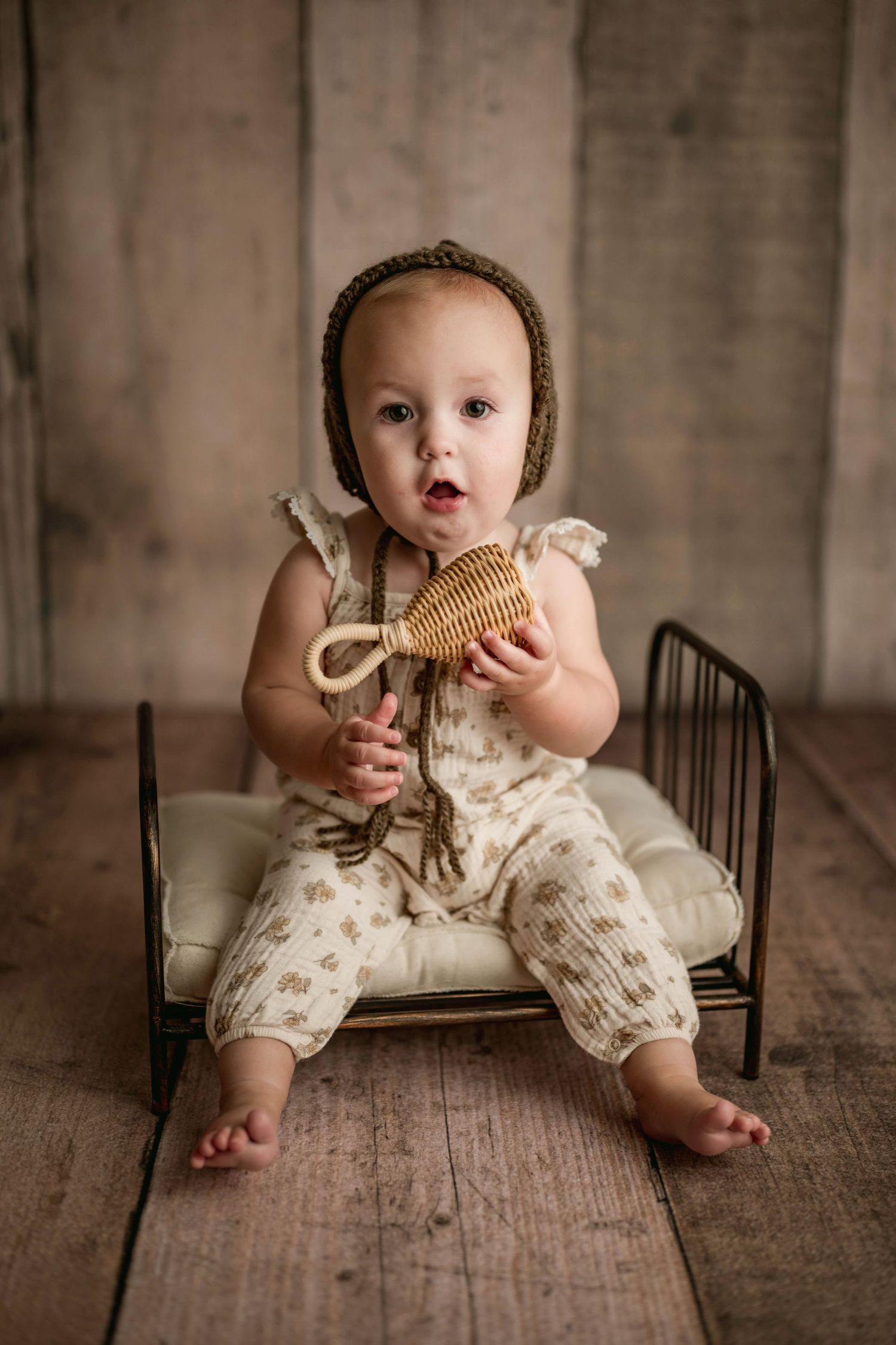 Curious baby in floral romper and brown bonnet holds wicker rattle while sitting on mini iron bed.
