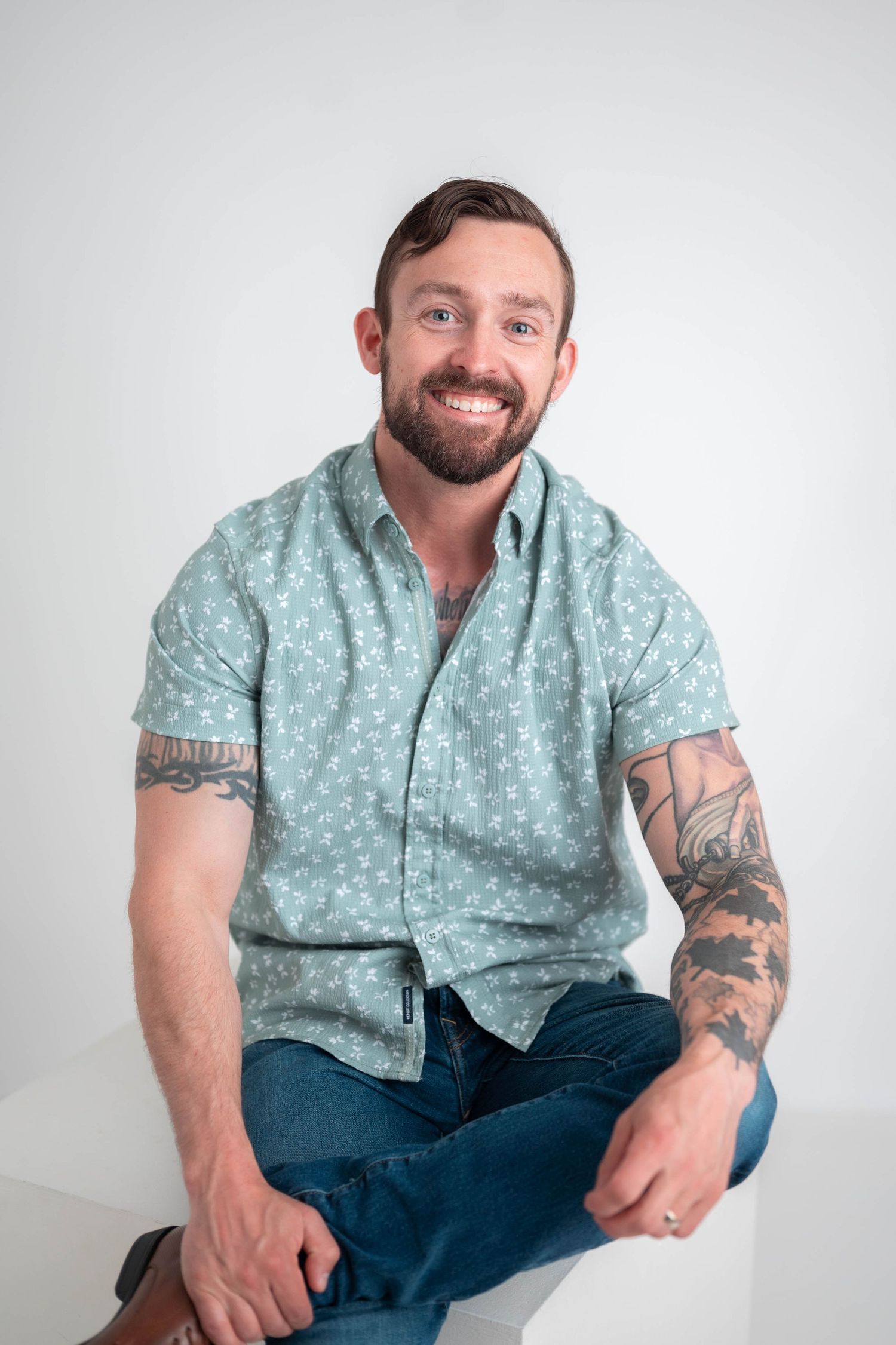Smiling bearded man with tattoos sits cross-legged wearing a light green floral button-up shirt and jeans.
