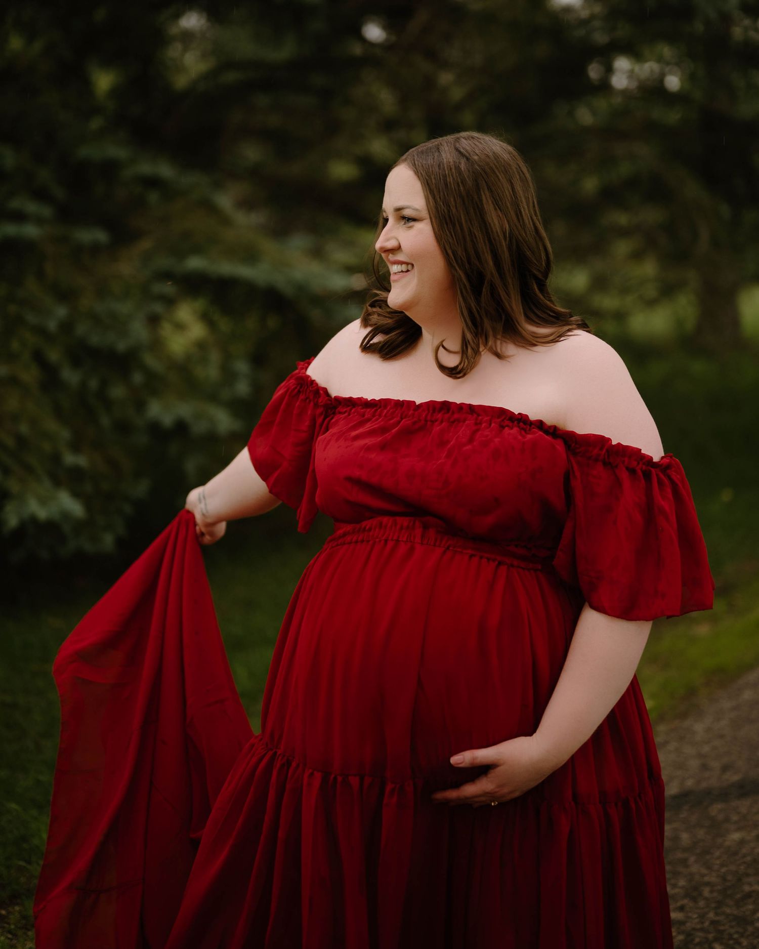 Pregnant woman in red off-shoulder maternity gown posing outdoors in lush green garden, smiling gracefully.