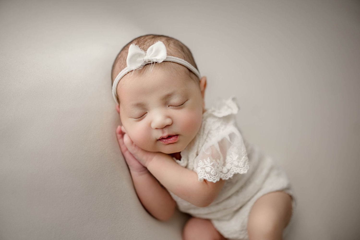 Sleeping newborn baby in white lace romper and bow headband posed on soft gray backdrop.