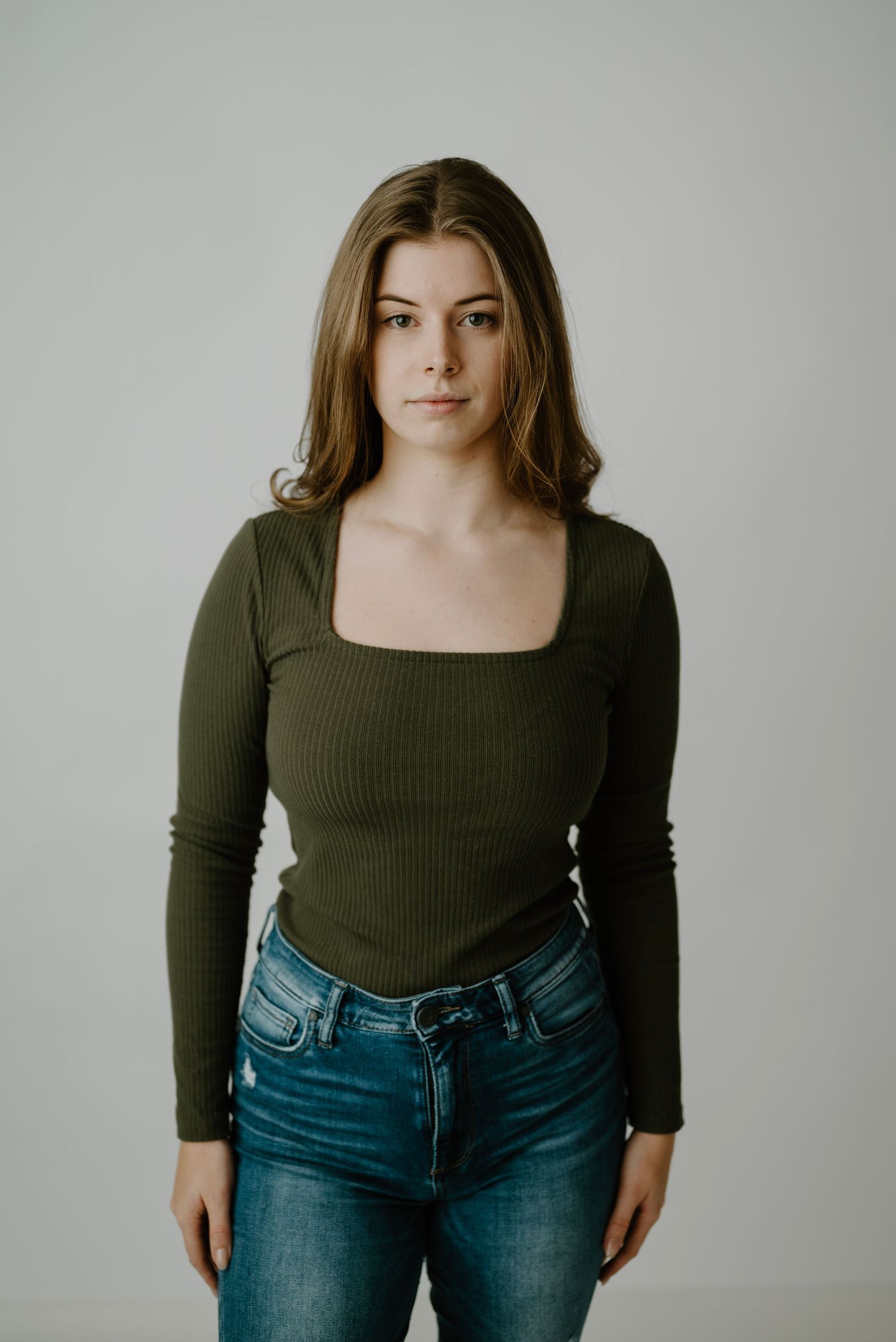 Young brunette woman in olive ribbed long-sleeve top and jeans standing against gray background.