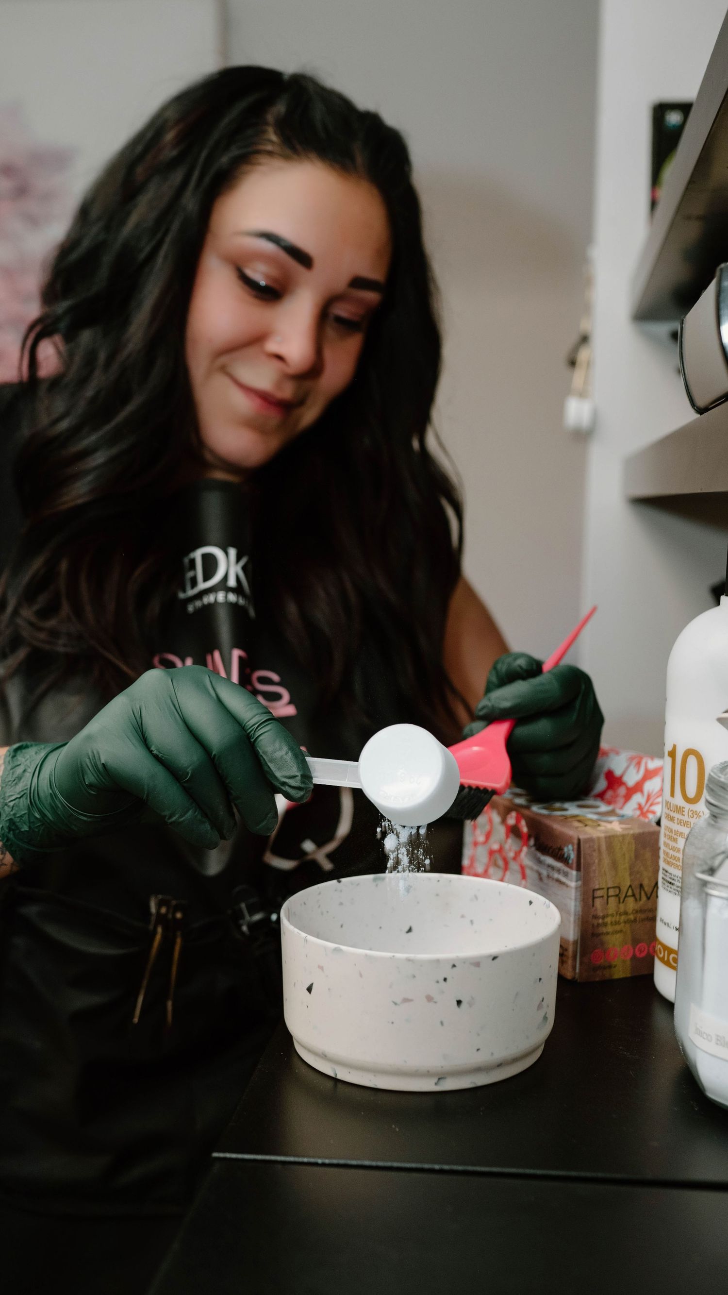 Female hairstylist wearing green gloves mixing hair color in bowl at salon station.