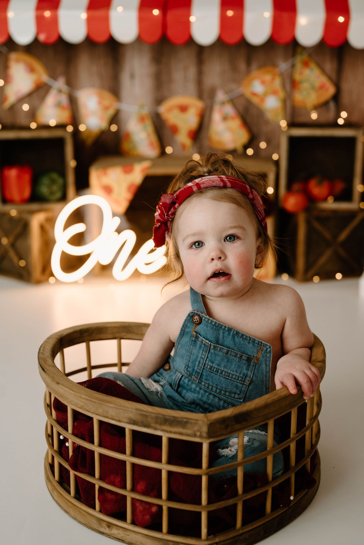 Baby girl in denim overalls sitting in wooden basket at pizza themed first birthday cake smash session.