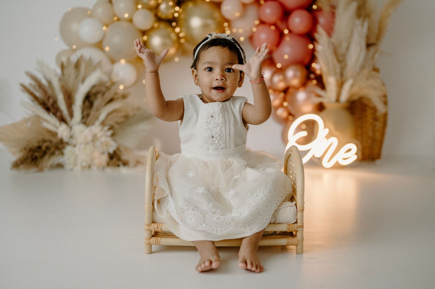 Baby girl in white dress sits on mini bed with balloon arch and neon One sign at first birthday photoshoot.
