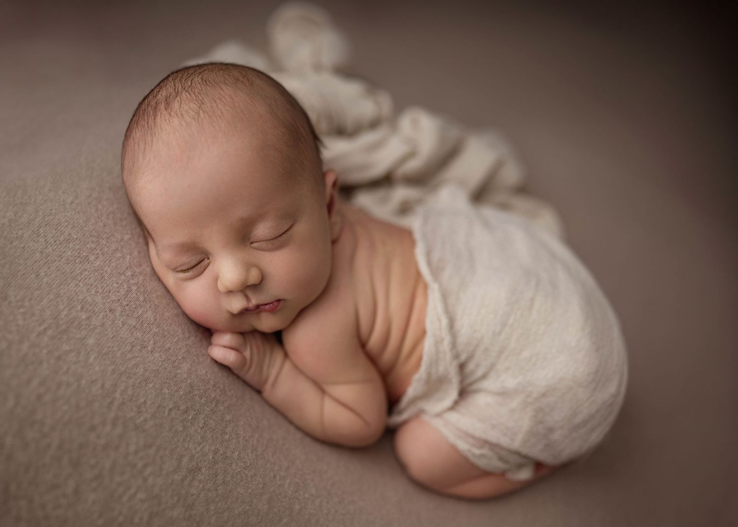 Peaceful newborn baby sleeping on a soft taupe blanket, wrapped in a cream swaddle during a professional newborn photo session.