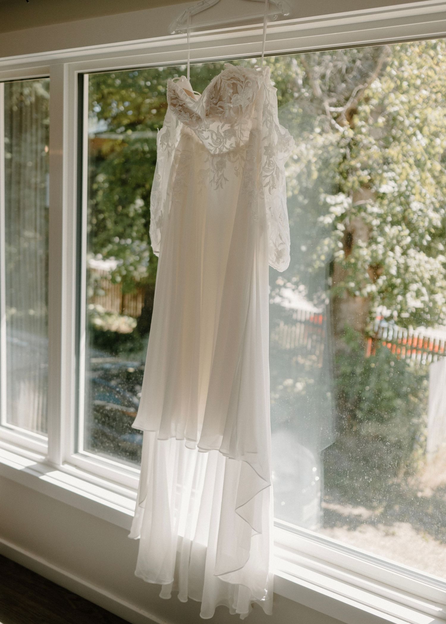 Elegant white lace wedding dress with long sleeves hanging in a sunlit window with trees visible outside.