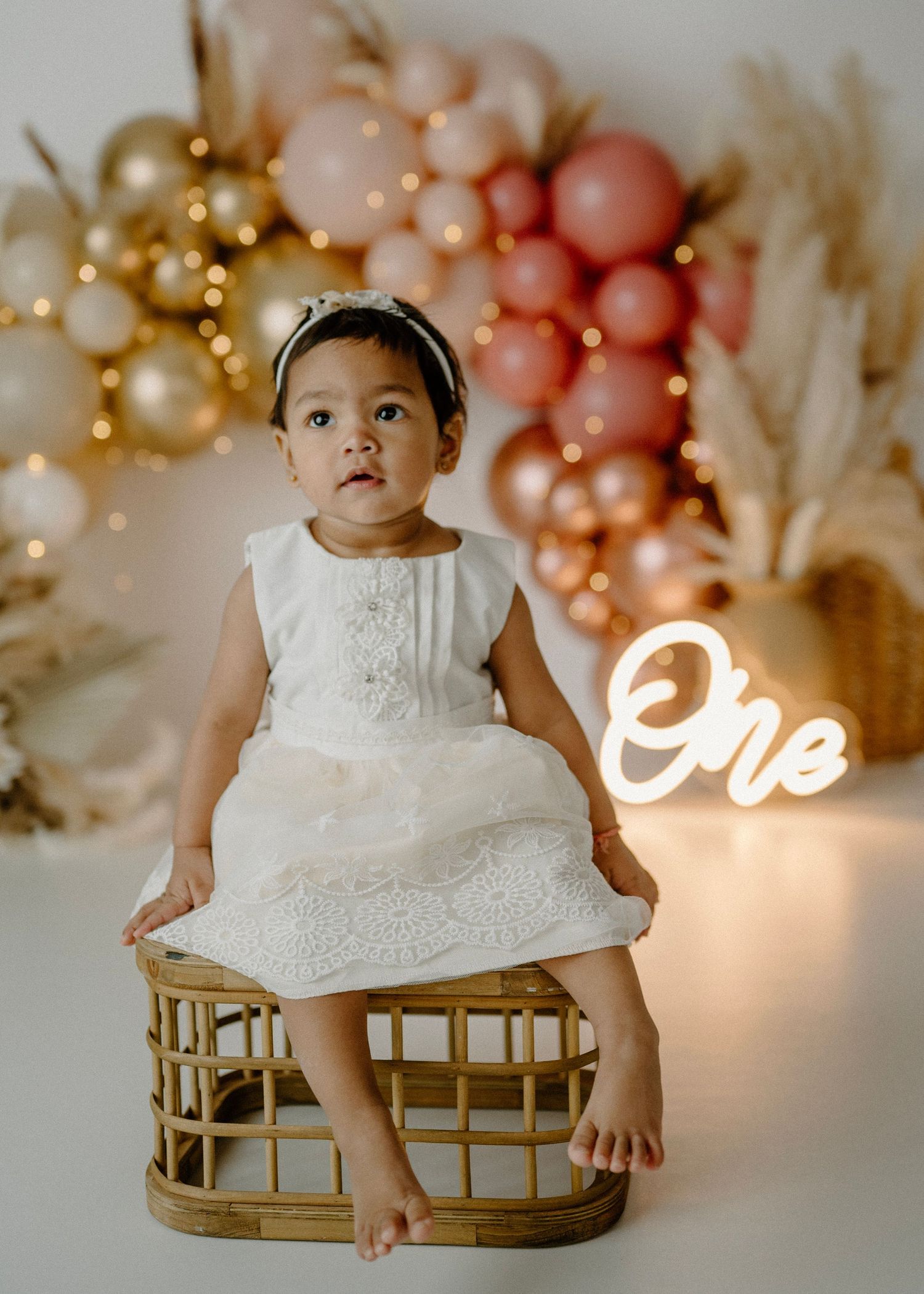 Baby girl in white lace dress sits on wicker basket at boho first birthday session with balloon garland.