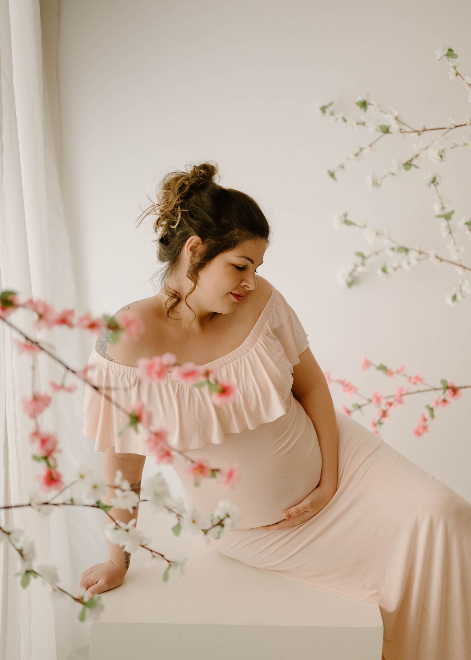Pregnant woman in blush ruffled off-shoulder maternity dress sits among delicate pink cherry blossom branches.