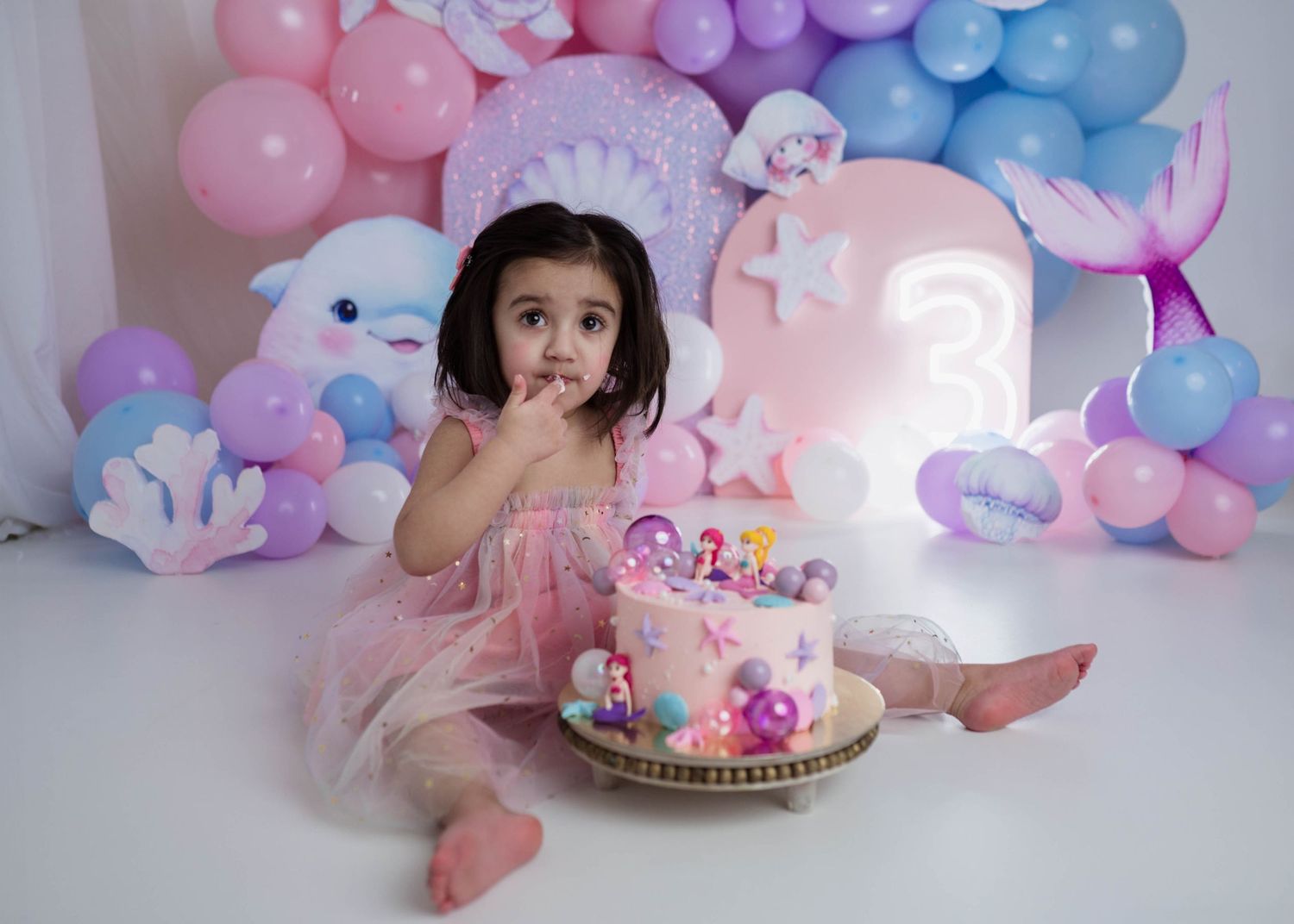Young girl in pink tulle dress sitting with mermaid birthday cake and colorful balloon backdrop.