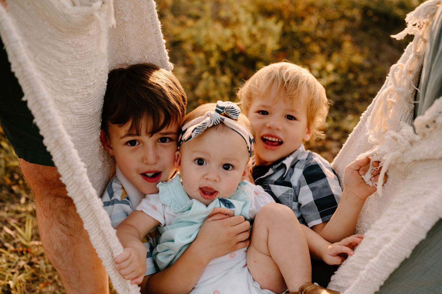 Three young siblings cuddle together in a cozy blanket hammock, smiling for a family portrait.