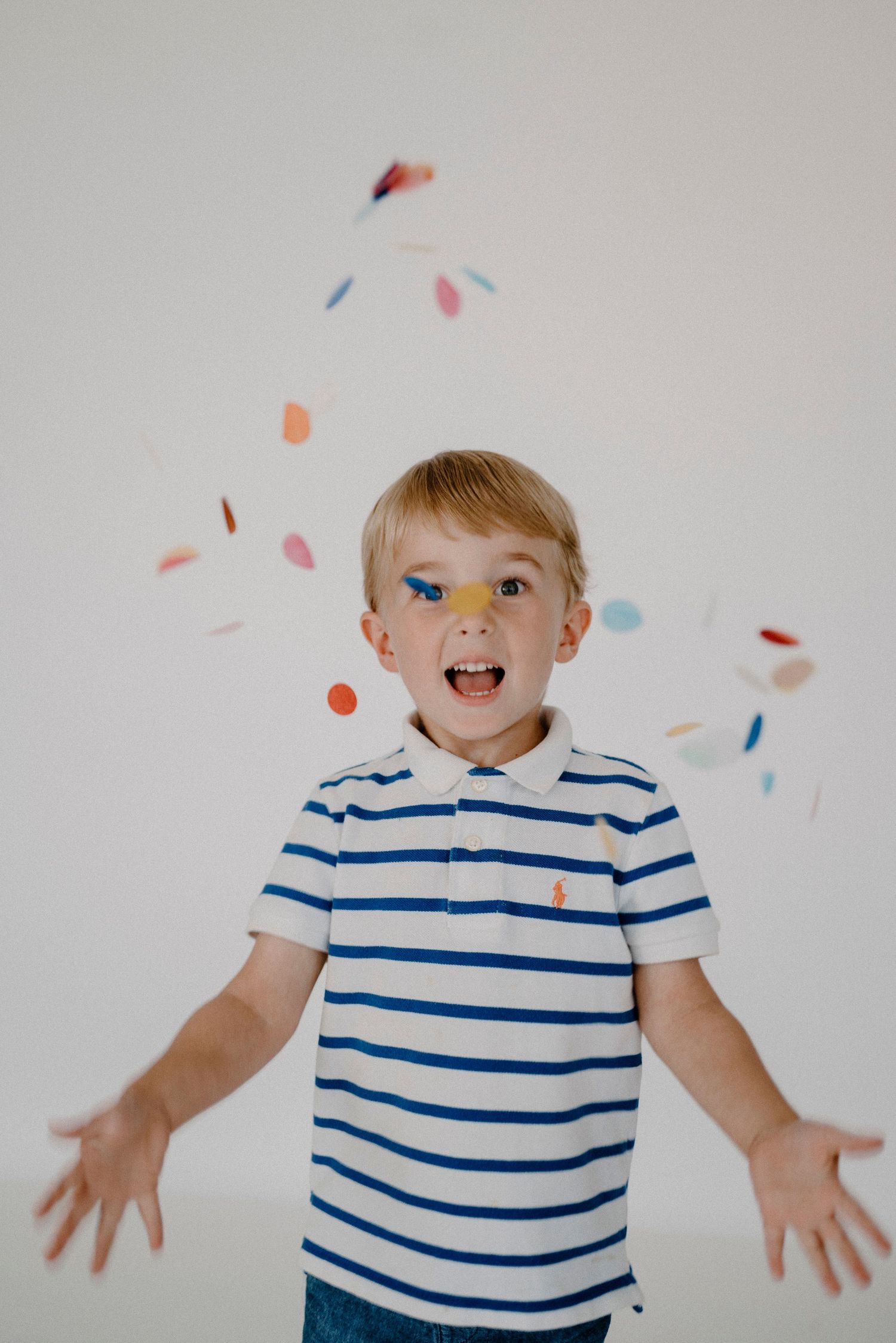 Enthusiastic blonde boy in striped polo shirt with confetti falling around him, arms outstretched.