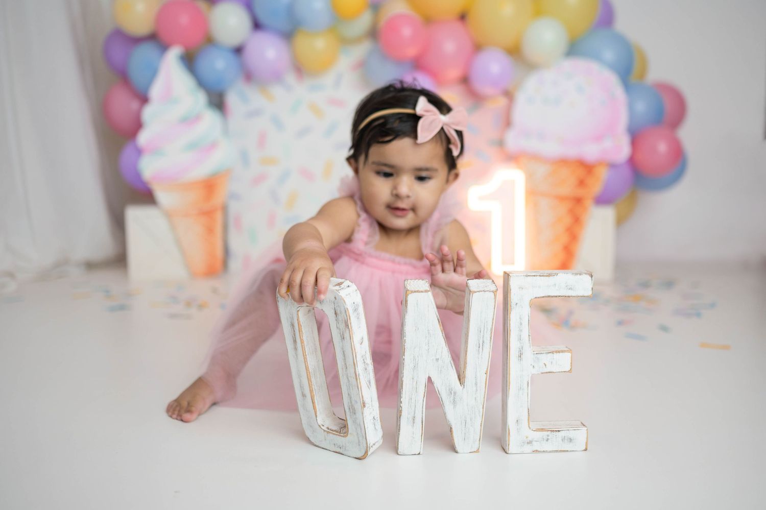 Baby girl in pink tutu poses with ONE letter sign at ice cream themed first birthday celebration.