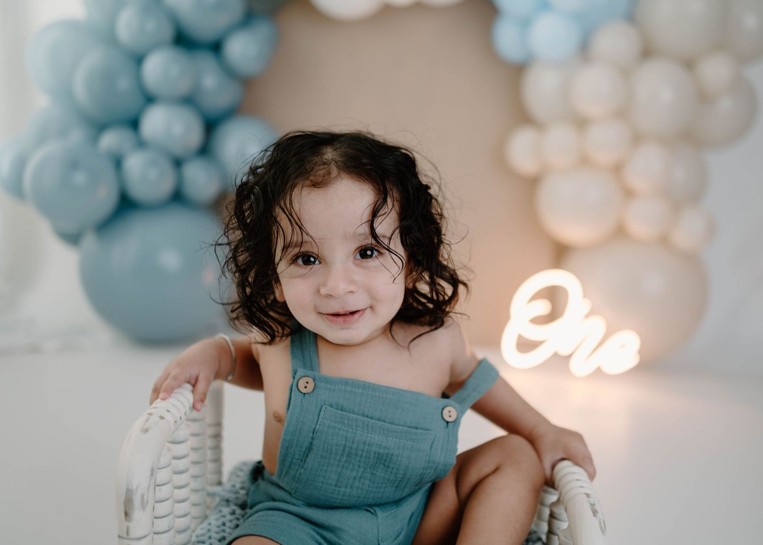 Cheerful toddler in teal romper sits in white wicker chair with blue balloon arch and glowing One neon sign.