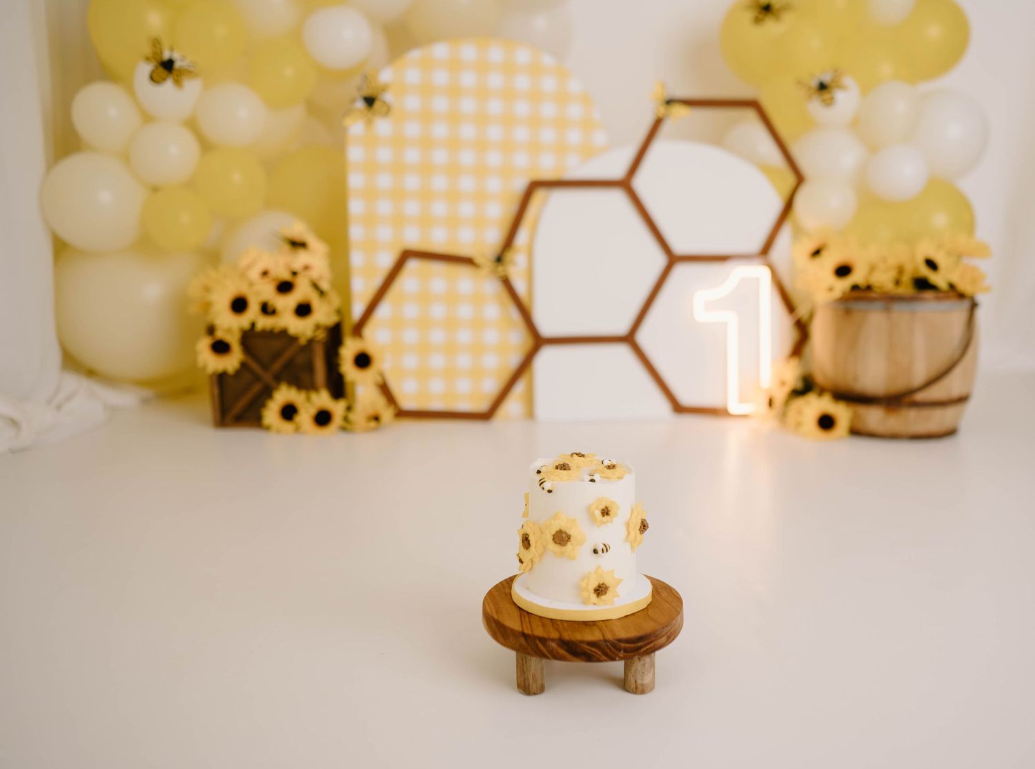 Bee-themed first birthday smash cake setup with honeycomb decor, sunflowers, yellow balloons, and a small white cake on a wooden stand.