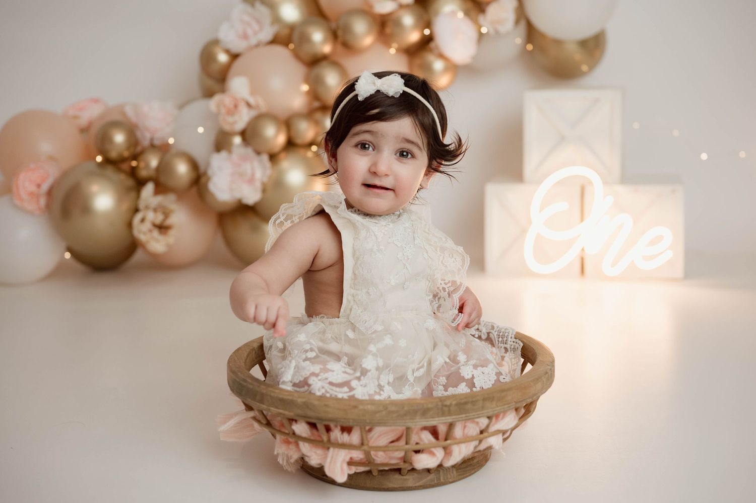 Baby girl in white lace dress sitting in wicker basket at elegant pink and gold first birthday cake smash.