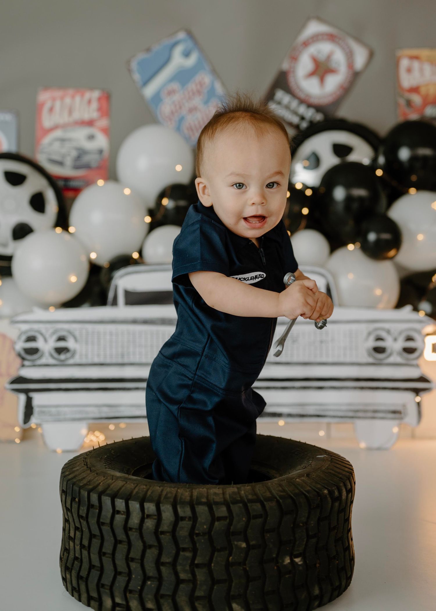 Smiling baby in mechanic jumpsuit stands on tire at automotive garage-themed first birthday smash.