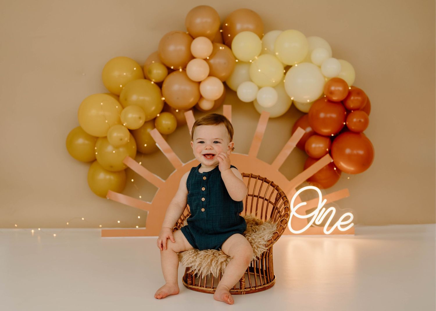 Cheerful baby boy in teal romper seated in wicker chair with colorful sun balloon arch.