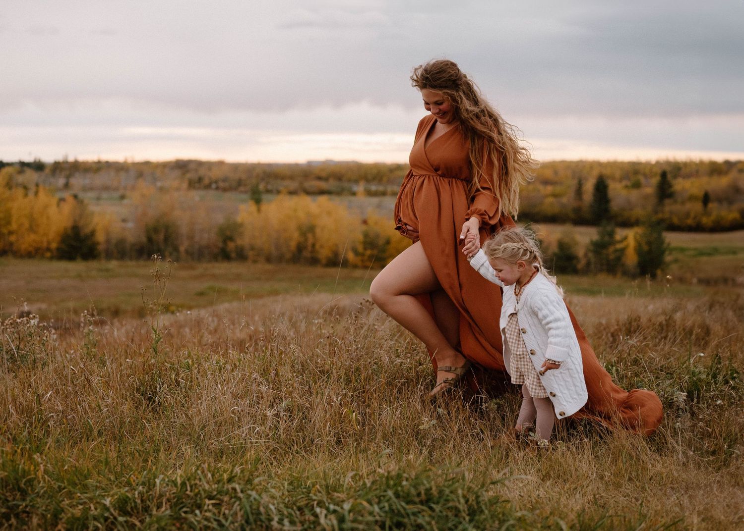 Pregnant woman in rust dress playing with young child in golden autumn field at dusk.