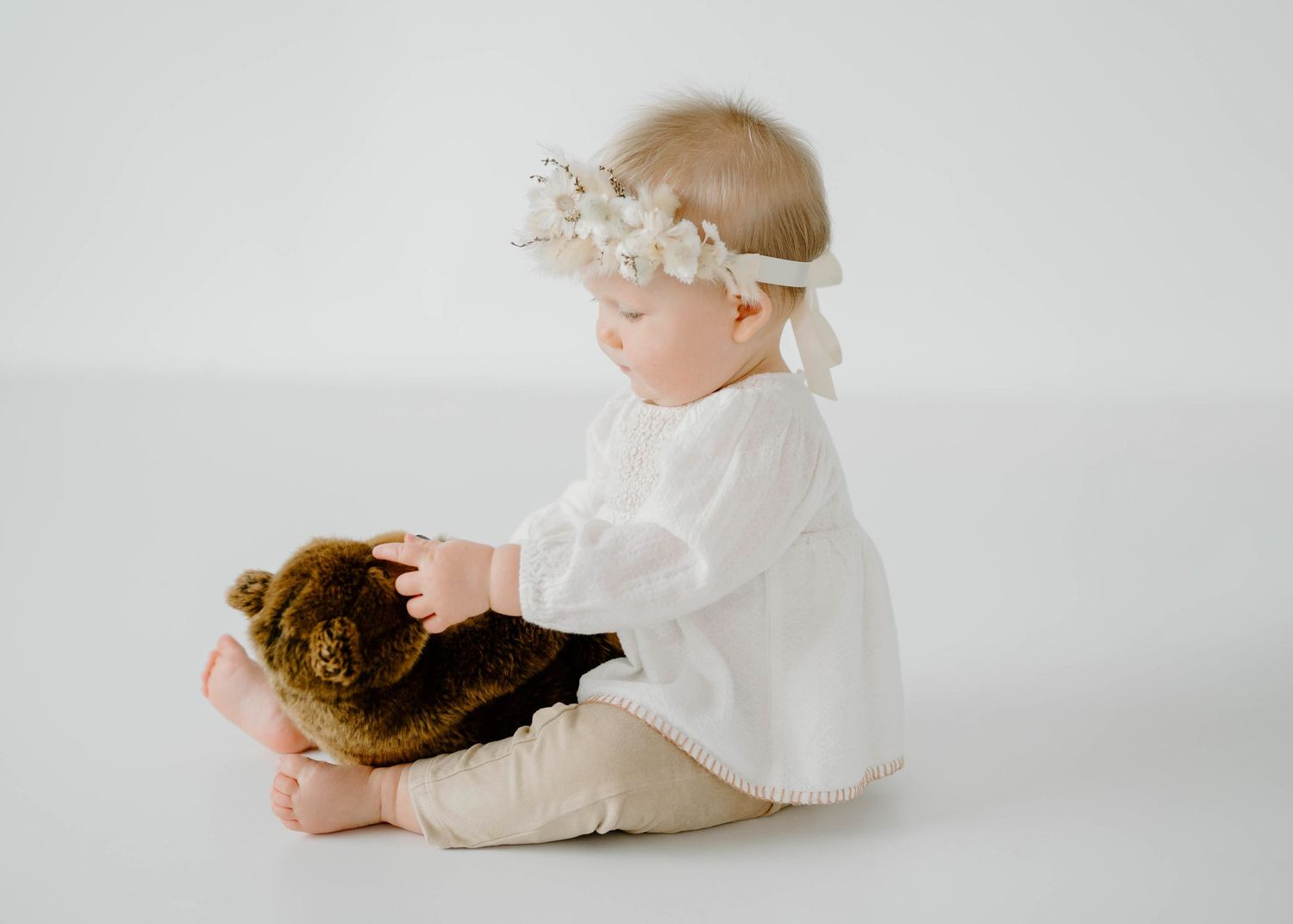 Cute baby in white outfit and floral headband sitting and holding a brown teddy bear.