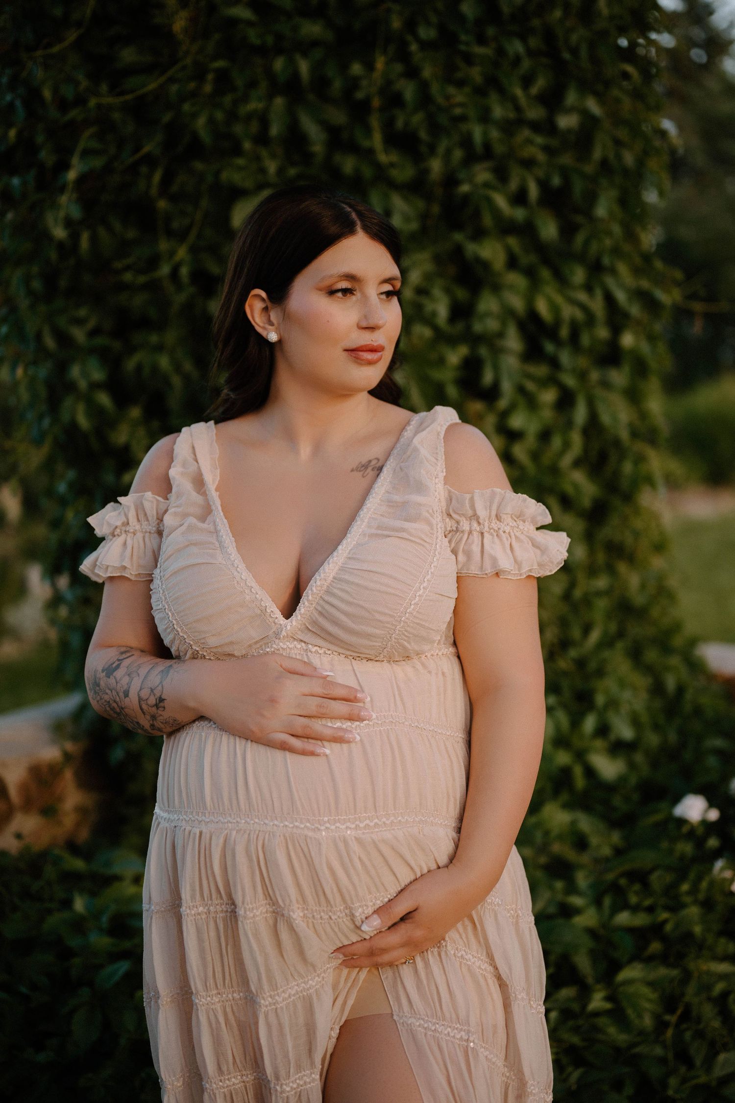 Pregnant woman in elegant cream dress cradling baby bump outdoors, surrounded by lush green foliage in golden light.