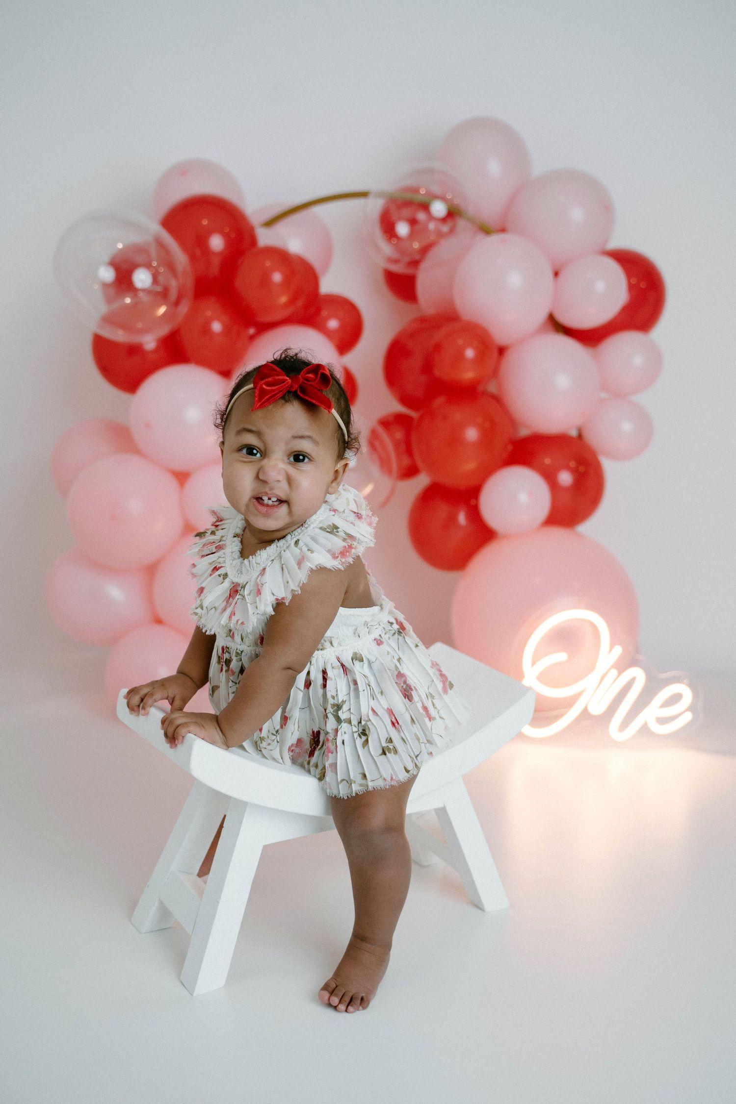 Smiling baby in floral dress stands at white chair with red pink balloon wreath and glowing One neon sign.
