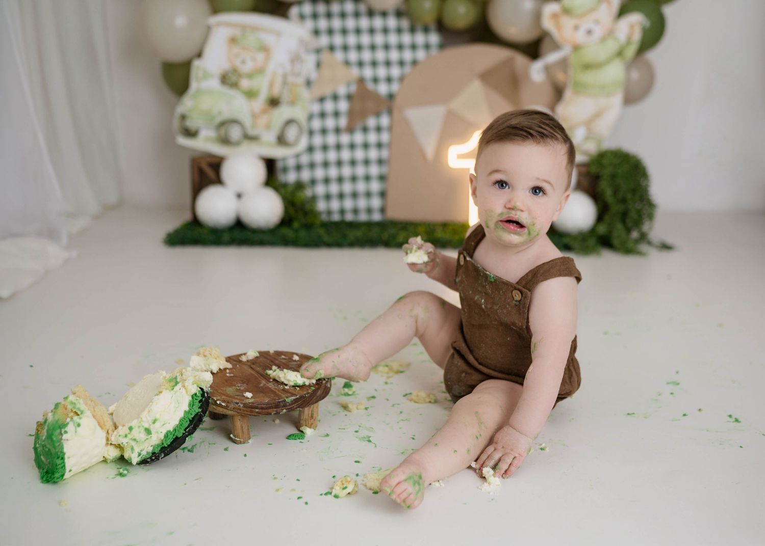 Baby boy in brown romper smashing green cake during first birthday cake smash photo session.