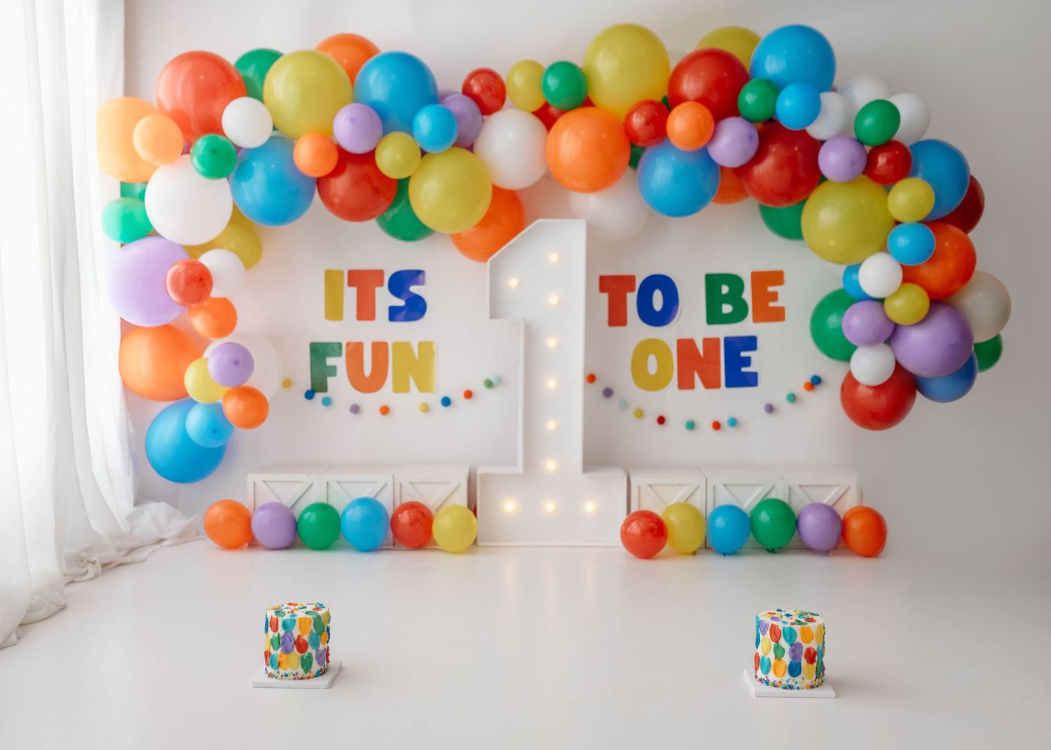 Colorful first birthday party backdrop with rainbow balloons and “It’s Fun To Be One” sign with smash cakes.