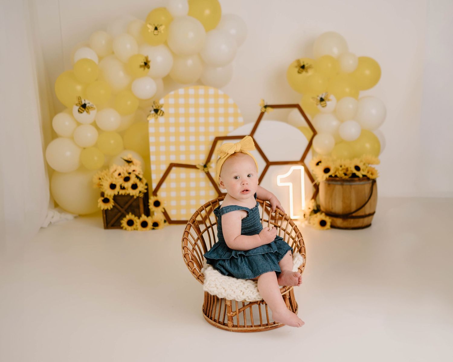 Baby girl in teal dress sits in wicker chair at bee-themed first birthday party with yellow balloon arch.