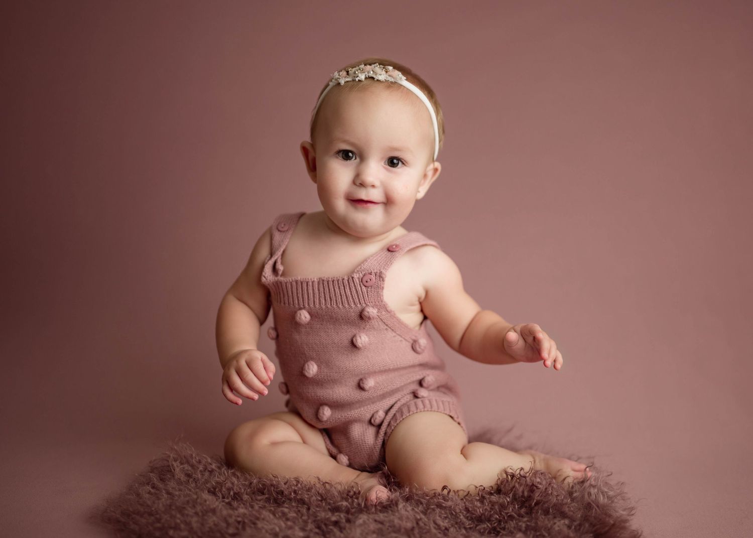 Happy baby girl in pink knit romper and floral headband sitting on fluffy fur rug.