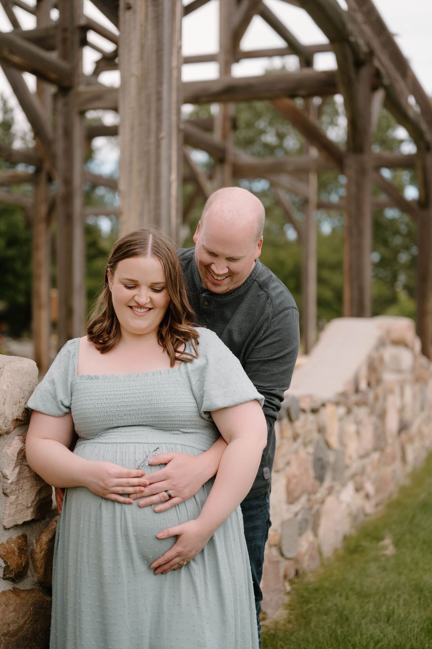 Laughing couple shares joyful maternity moment outdoors, woman in sage dress holding baby bump near wooden pergola and stone wall.