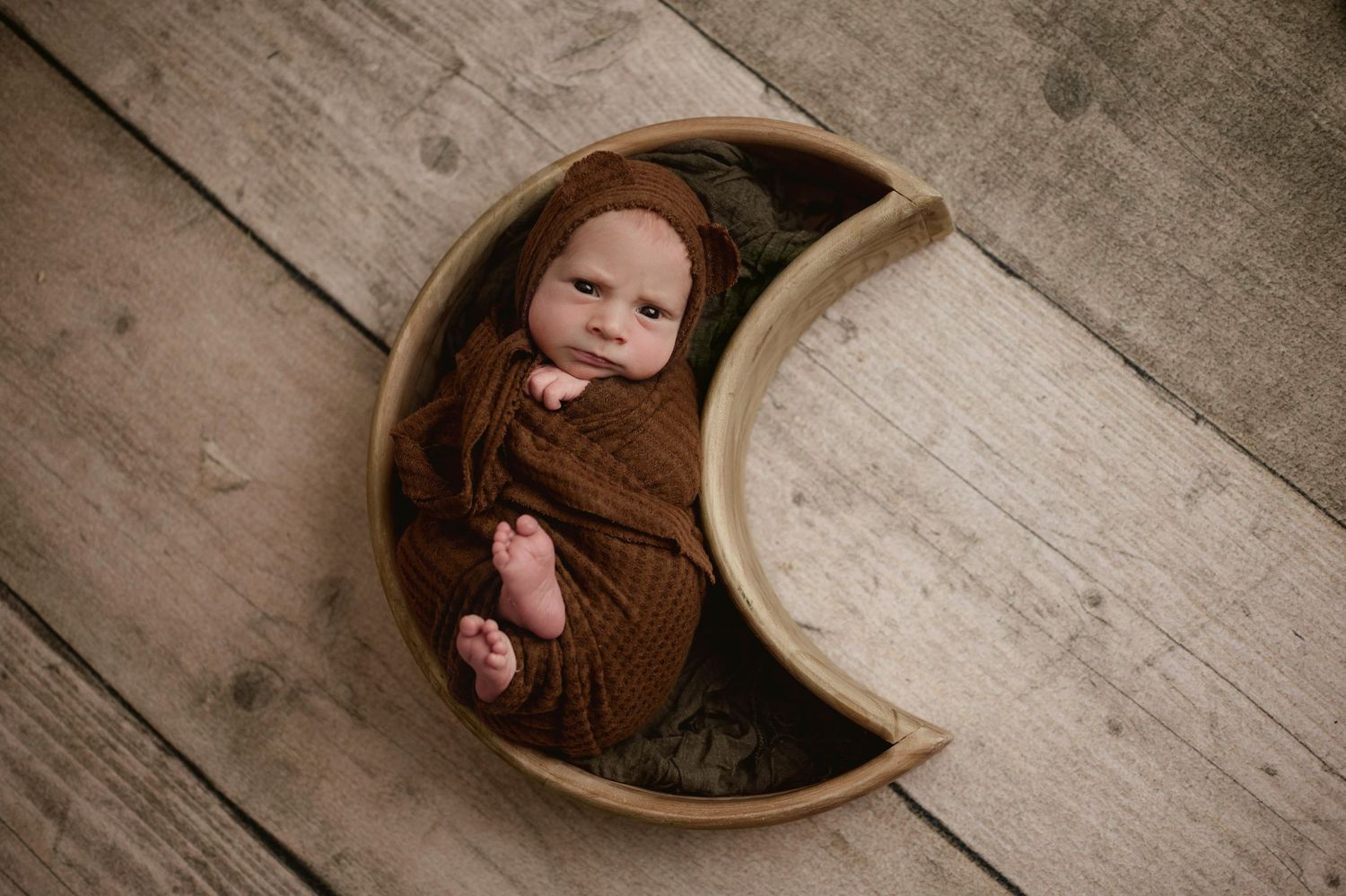 Smiling newborn baby wrapped in brown swaddle lying in a crescent moon-shaped wooden bowl.