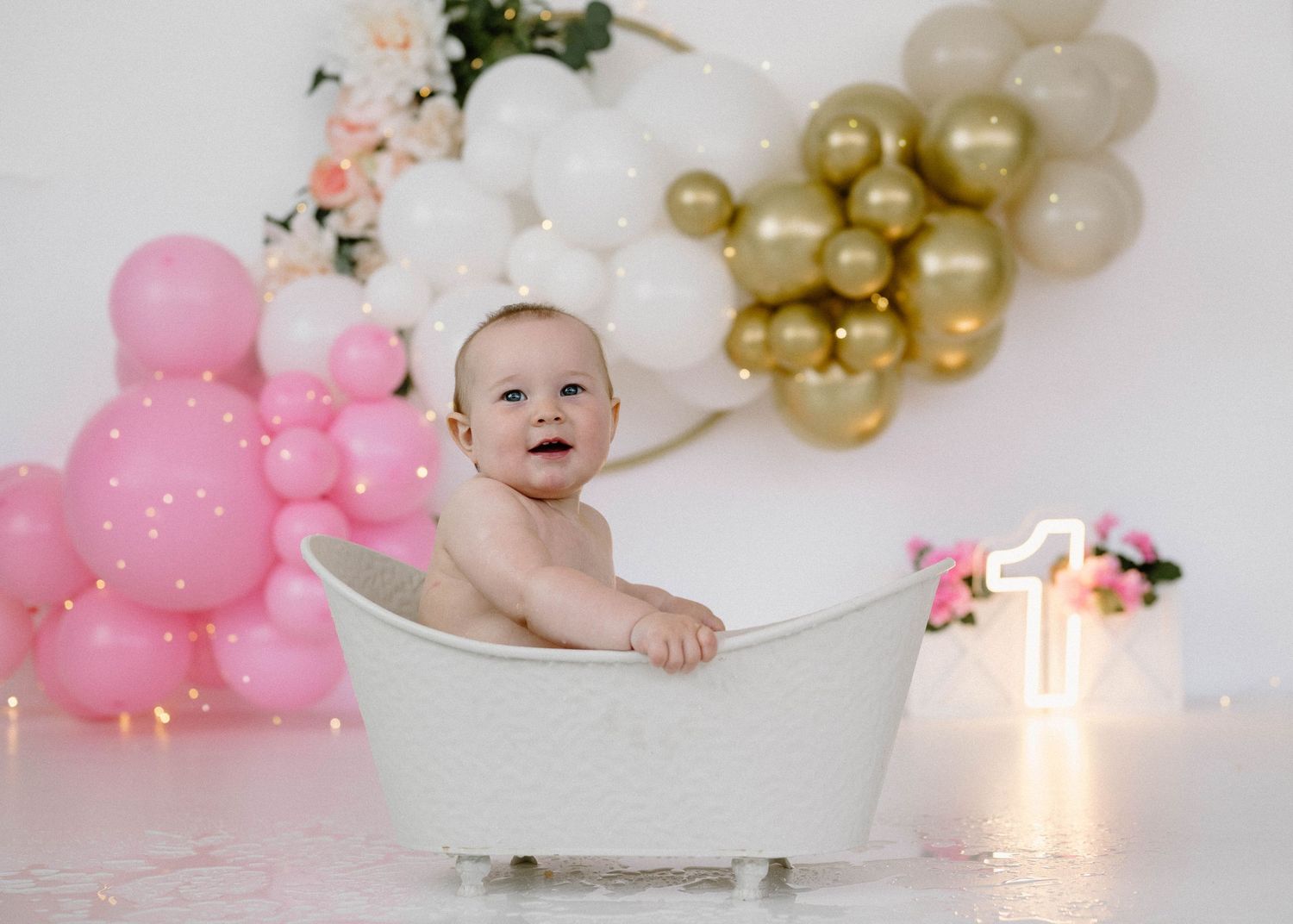 Adorable baby sits in small white bathtub surrounded by pink, white, and gold balloon garland decor.