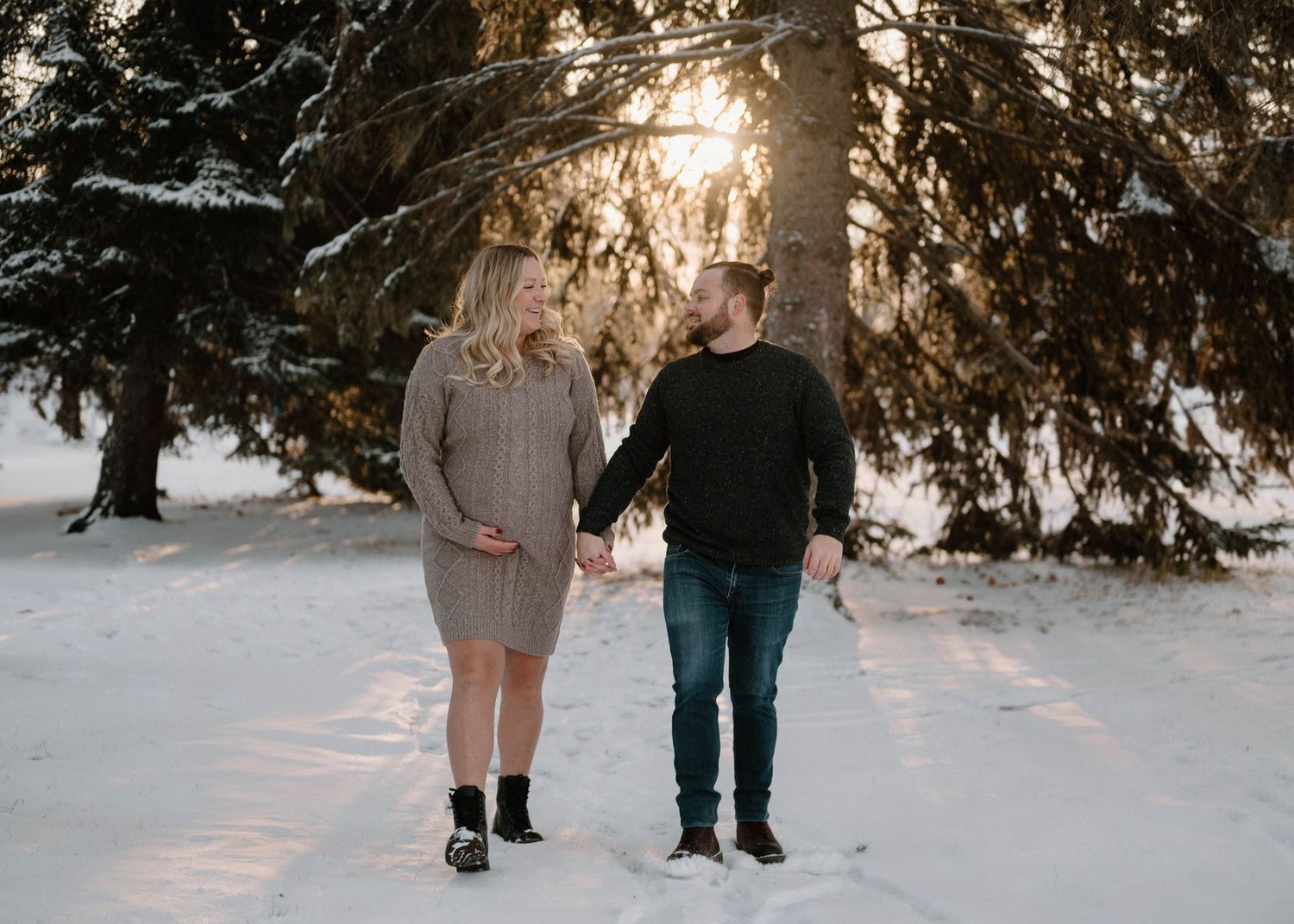 Expecting couple holding hands walking through snowy pine forest At Fred Johns Park during golden hour maternity shoot.