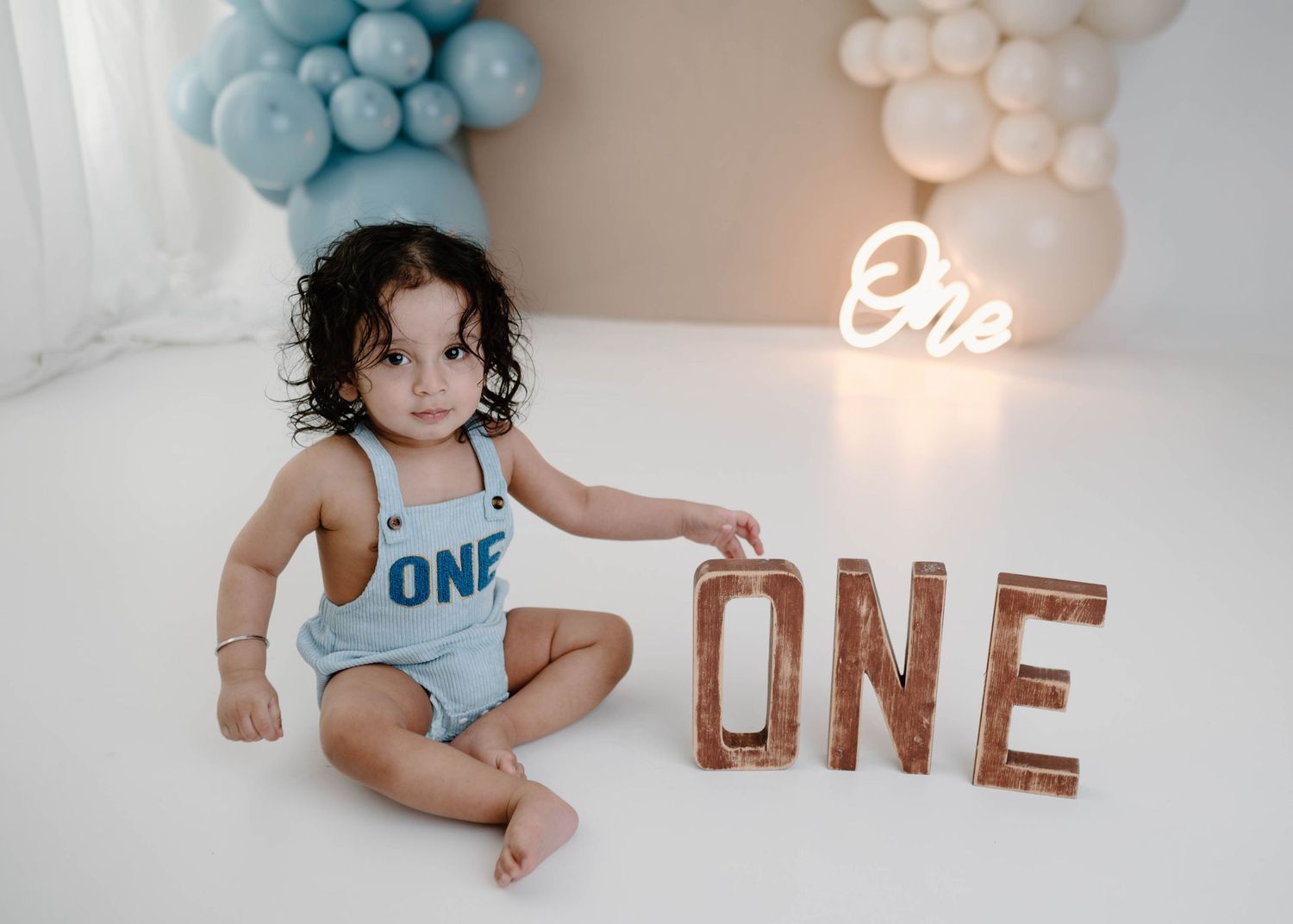 A toddler in a blue ONE romper sits beside wooden ONE letters with a neon sign and balloon arch.