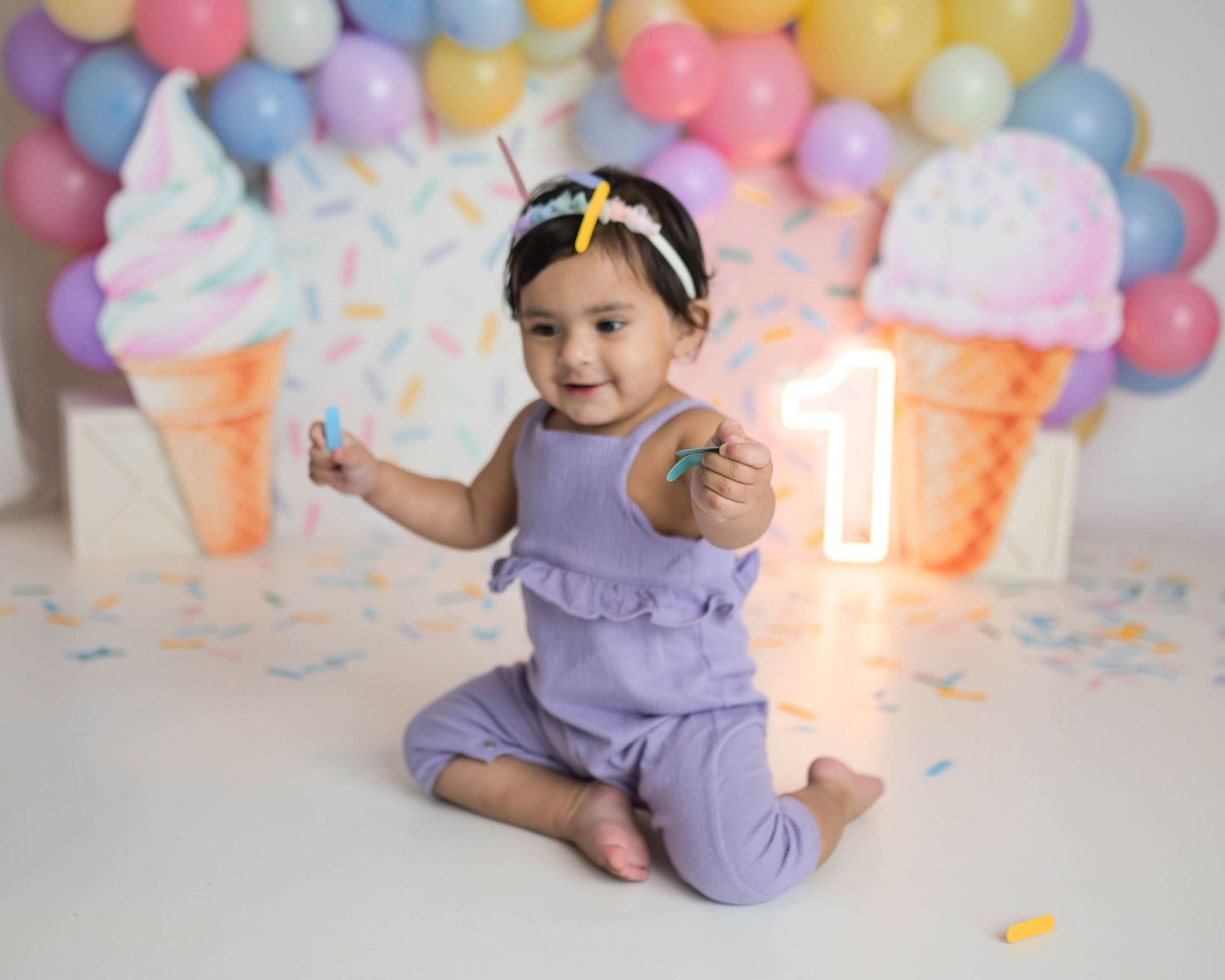 Baby girl in purple outfit plays with confetti at ice cream themed first birthday with colorful balloons.